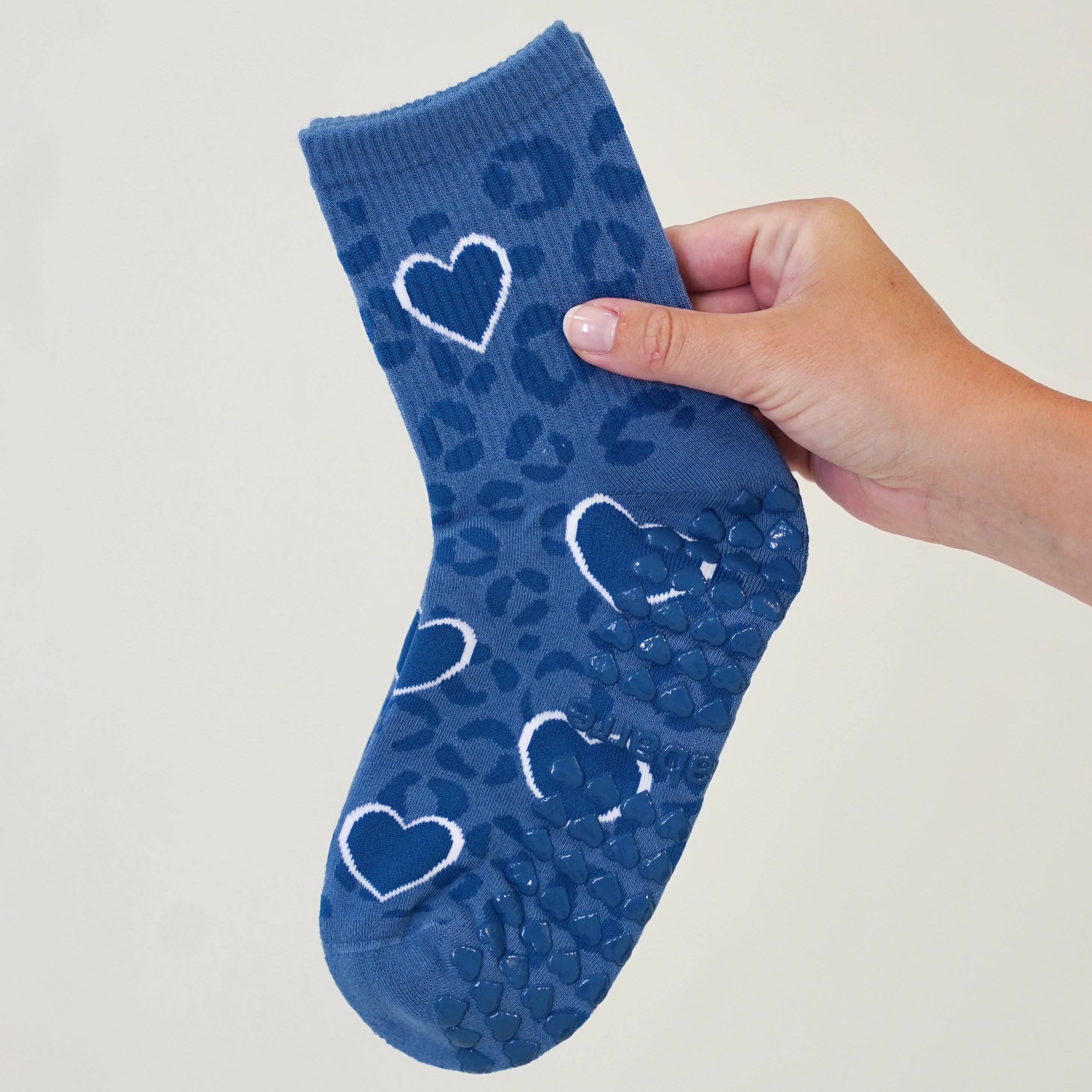 A blue ankle sock with a leopard print pattern and white heart outlines, held up by a person's hand against a plain light background. Cheetah Grip Socks