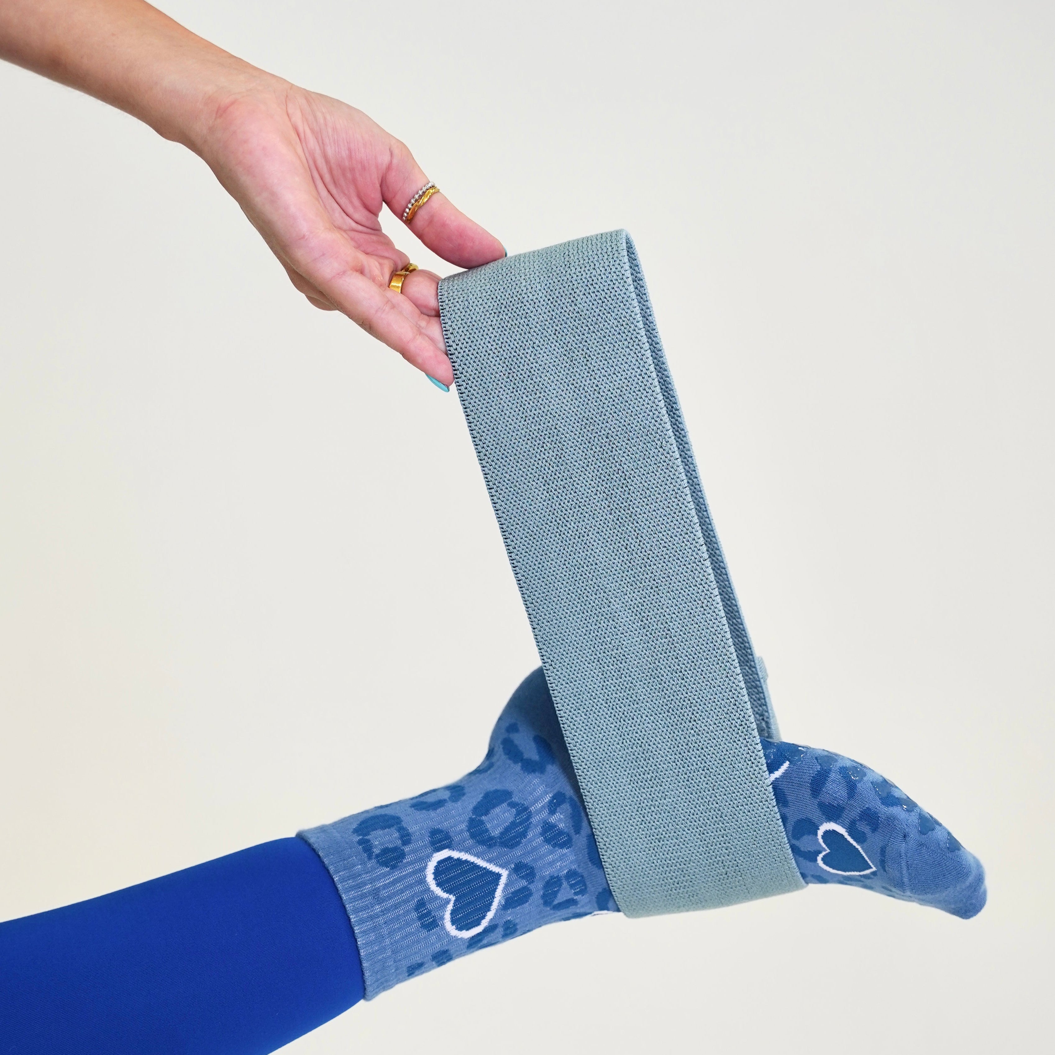 A person's foot and leg extended upward while wearing blue patterned socks with white heart details and a solid blue tights leg, with a hand holding a gray fabric strap around the ankle. Cheetah barre Socks- blue