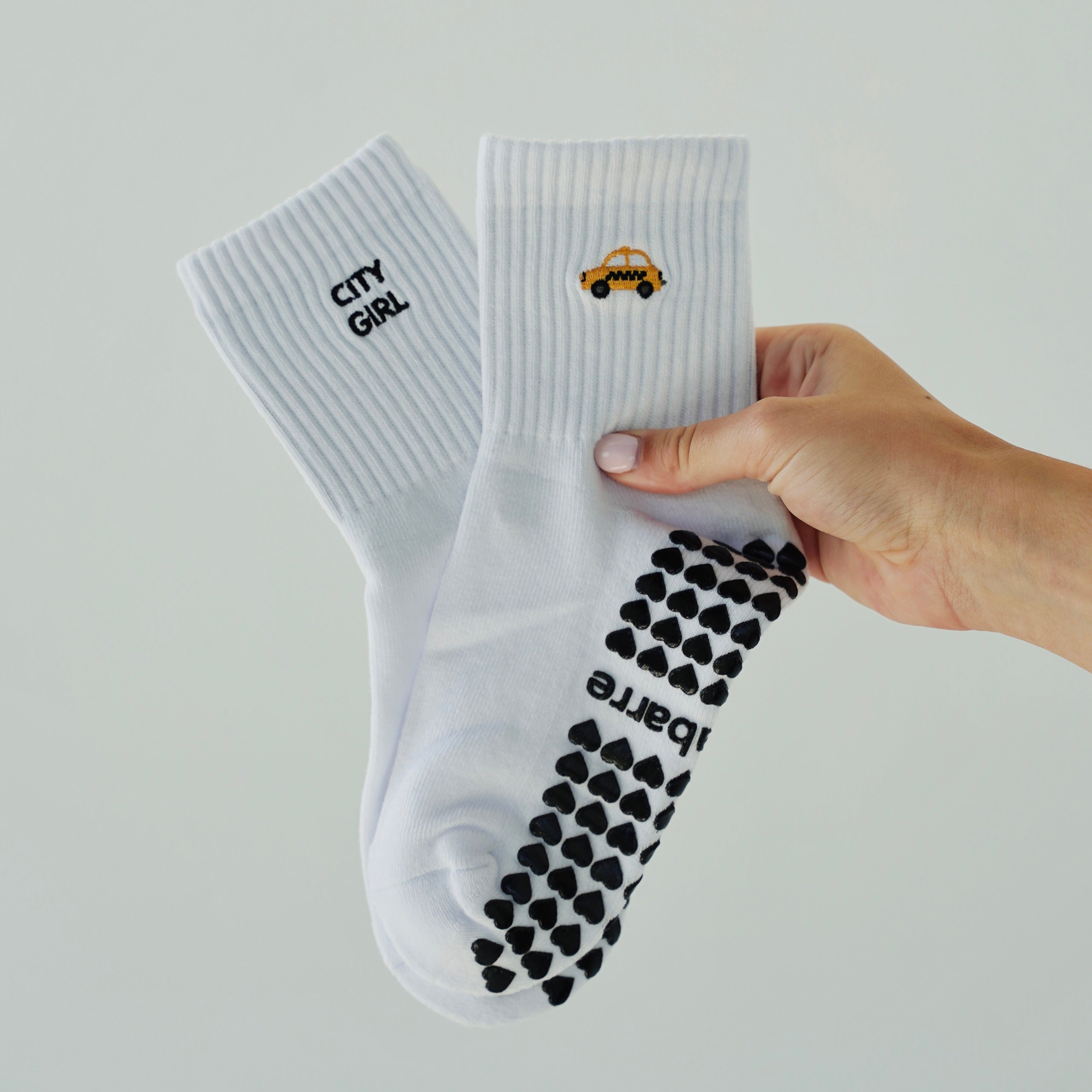 A hand holding a pair of white grip socks against a light gray background. One sock has black text reading 'CITY GIRL' and a small taxi, while the other shows a black grip pattern on the sole and the brand name 'Pilabarre'.