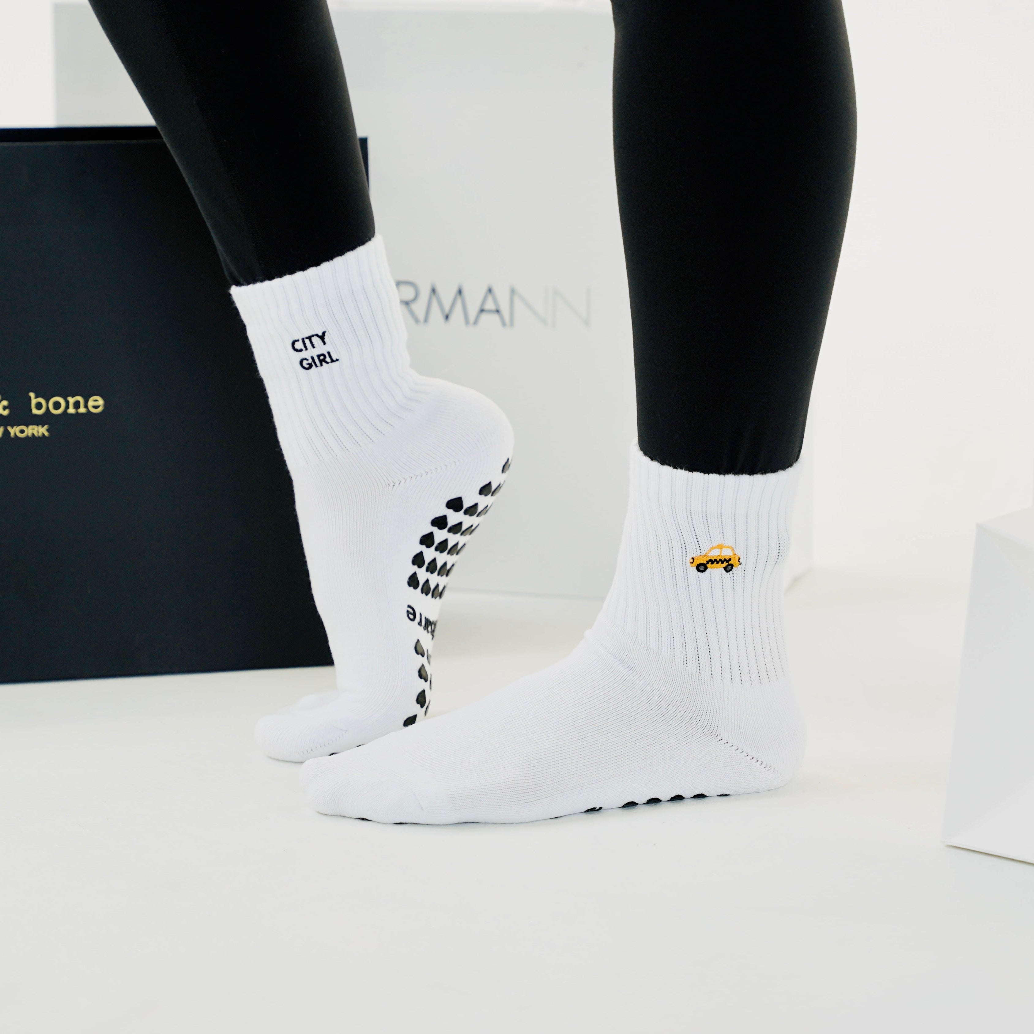 White grip socks worn on a person's feet, shown with black leggings. The socks feature black grip dots on the soles and a small yellow taxi on the side.
