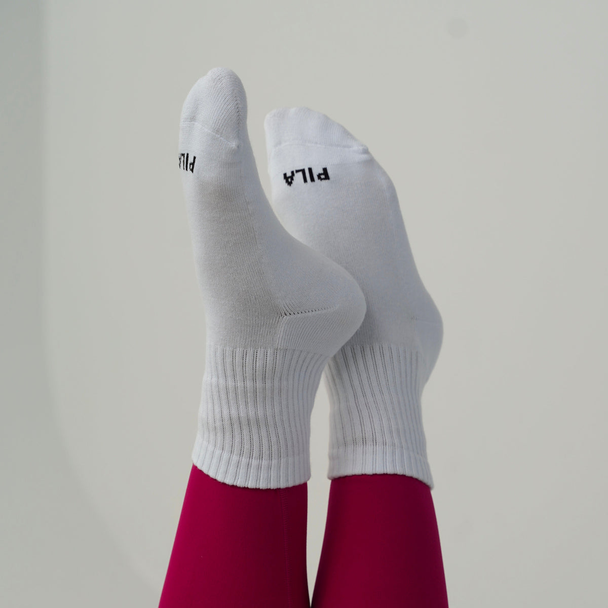 White athletic crew socks with black PILA branding, worn on legs covered in magenta tights, photographed against a plain light gray background.