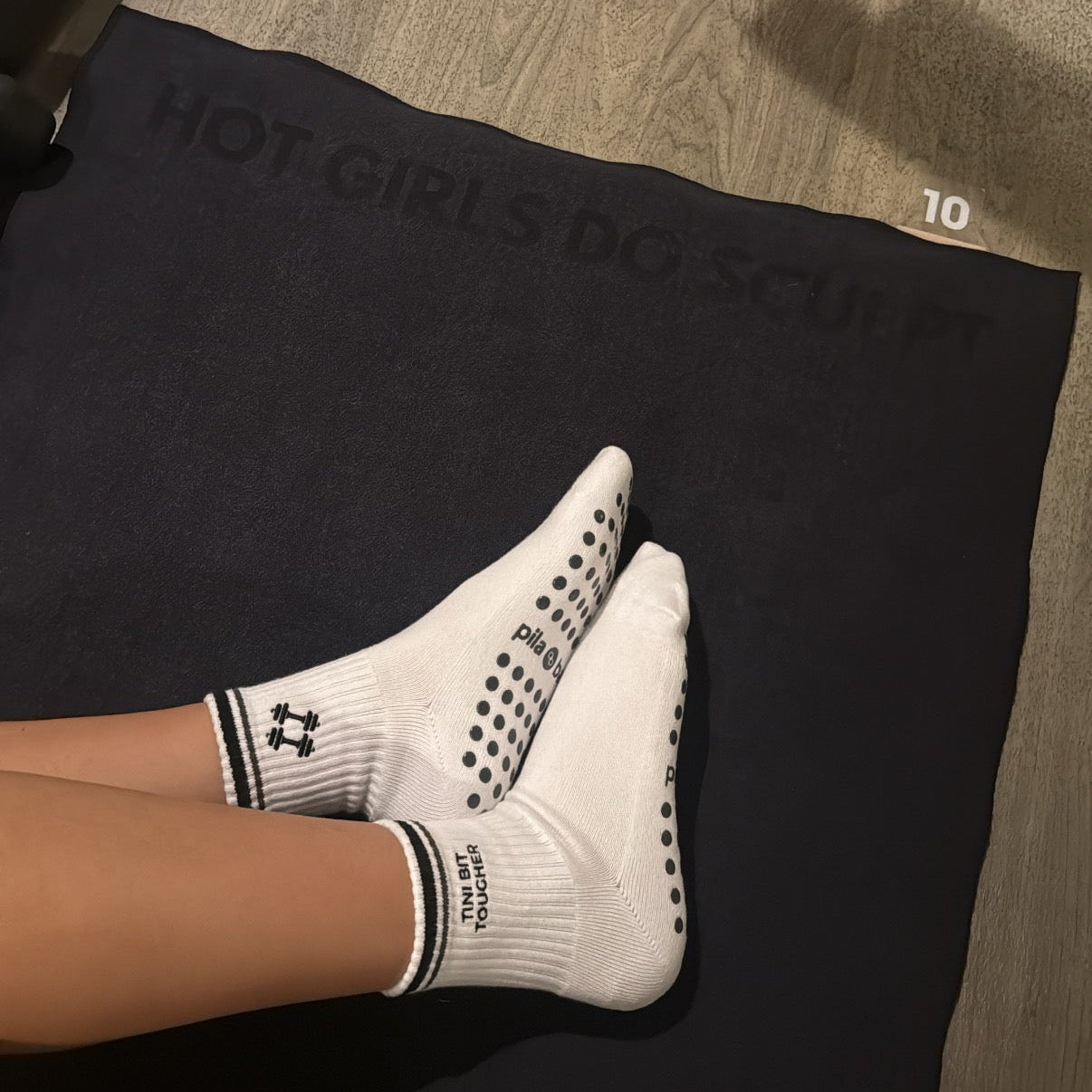 White grip socks with black dots and stripes worn on feet resting on a black yoga mat on a wood floor.