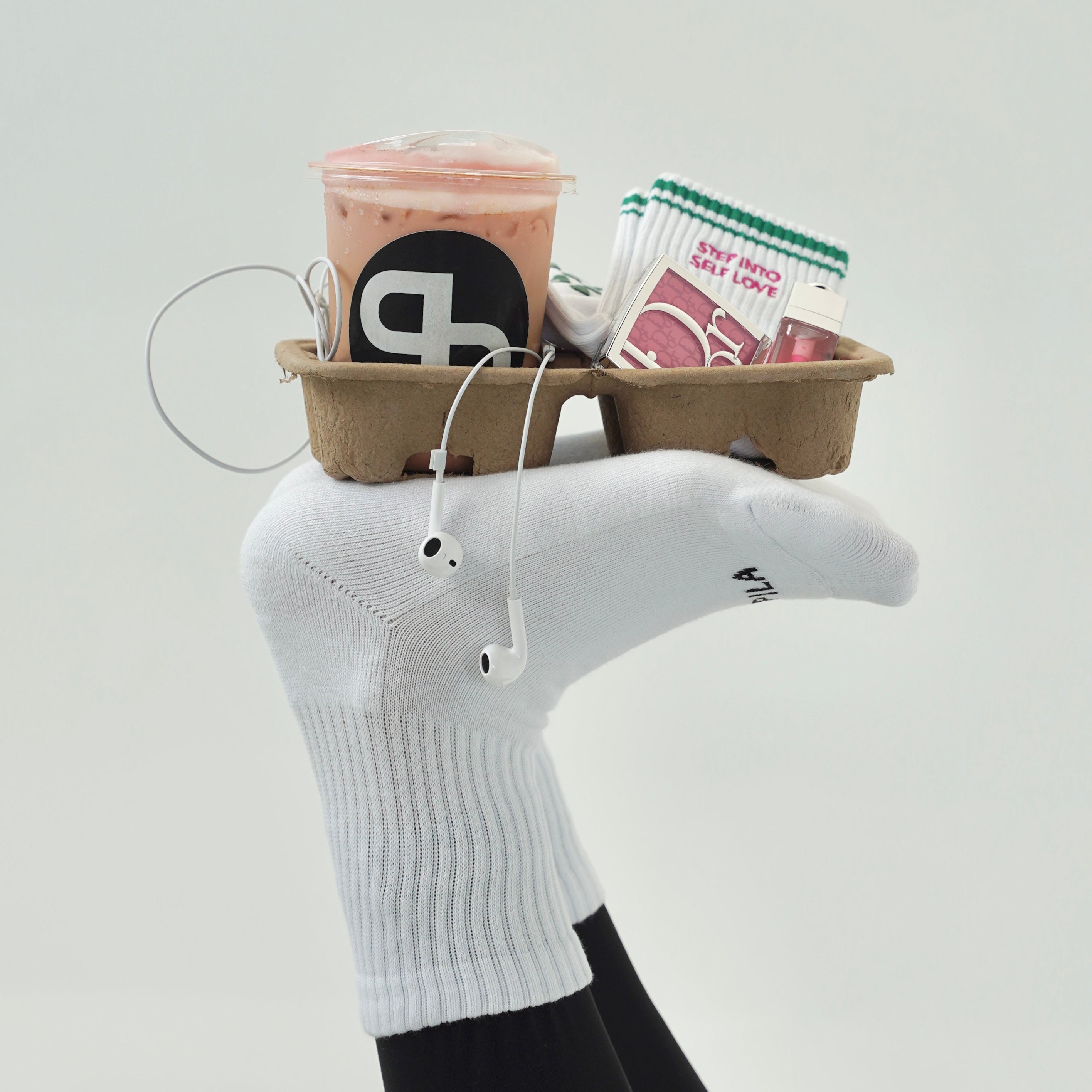 A white ribbed crew sock worn on a foot, holding a small brown paper tray with a pink drink, earbud wire, and a small packaged item. The foot is also wearing black tights against a plain light gray studio background.