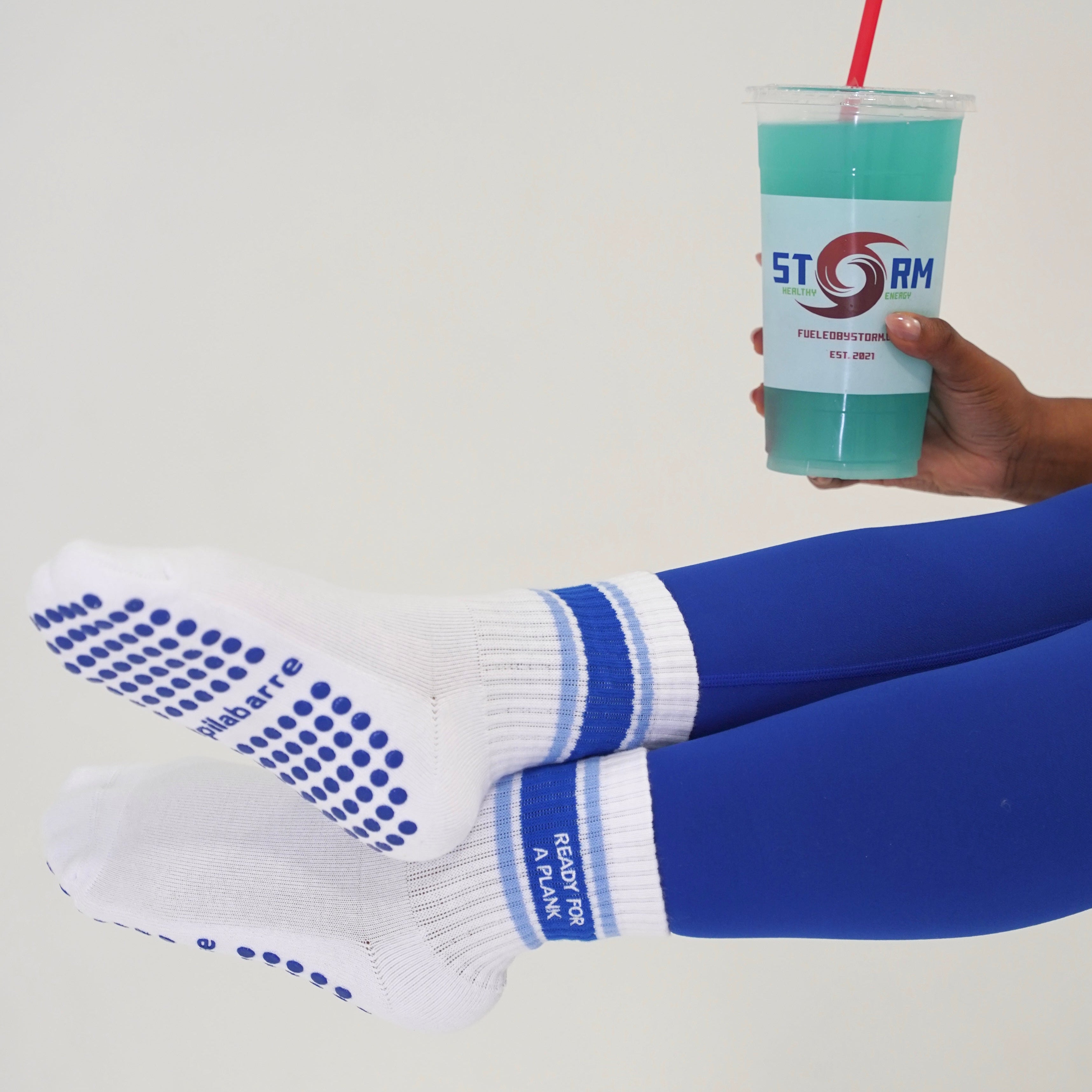 White grip socks with blue dots on the soles and blue stripes on the cuffs, worn by a person sitting with their legs raised, holding a blue drink with a red straw. The person is wearing blue leggings against a plain white background. Ready for a plank barre socks
