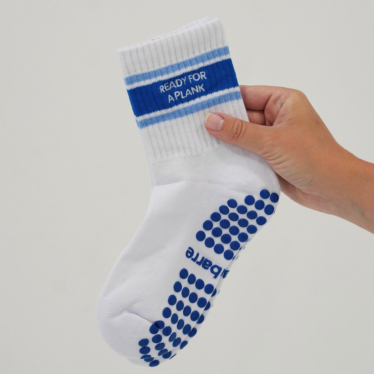 A hand holding a white baby sock with blue grip dots on the sole and blue stripes on the cuff that read 'READY FOR A PLANK'. Ready for a plank grip socks