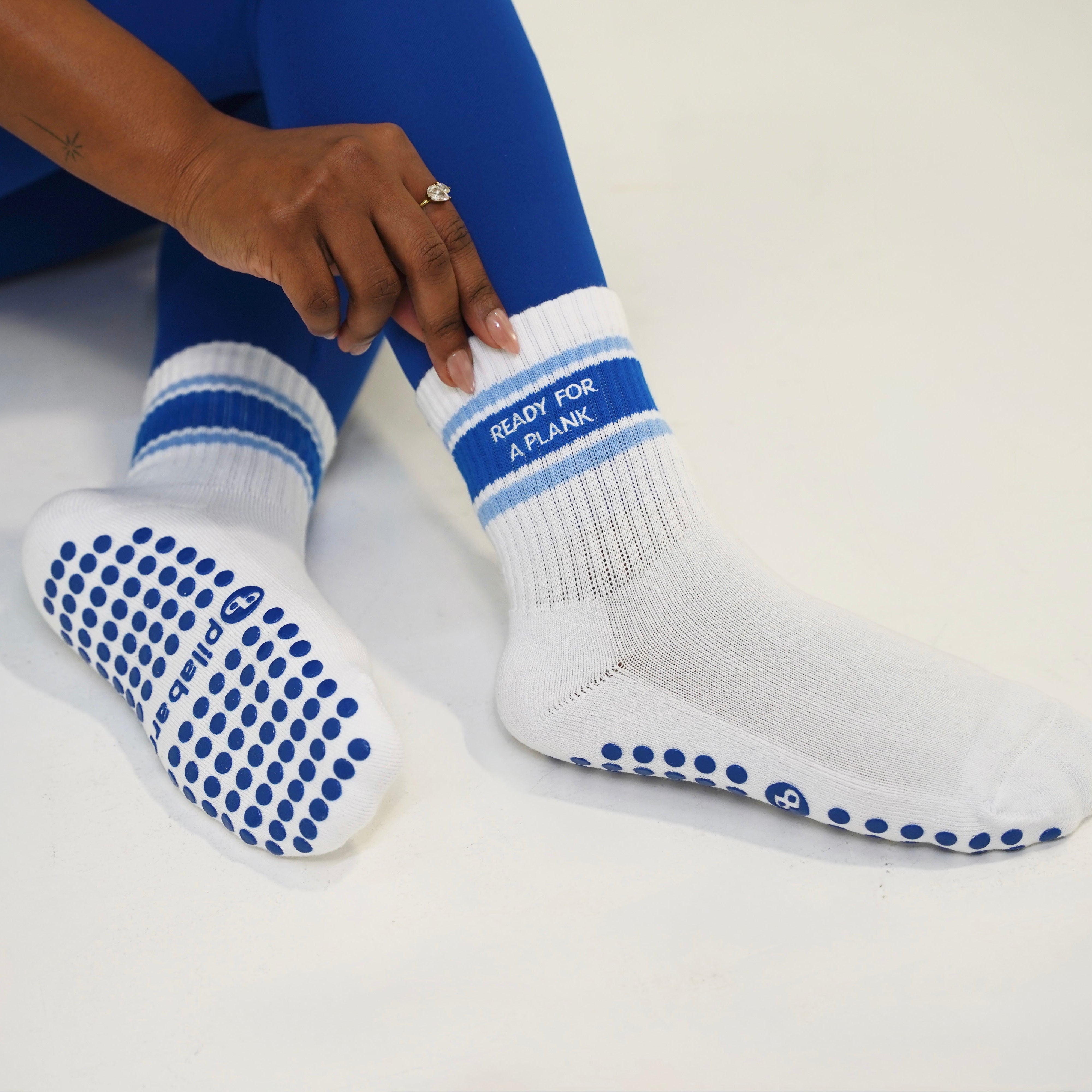 White grip socks with blue stripes and blue silicone dots, shown on a person's feet and lower legs against a light studio background. One sock has the text 'READY FOR A PLANK' printed on it. Ready for a plank pilates socks
