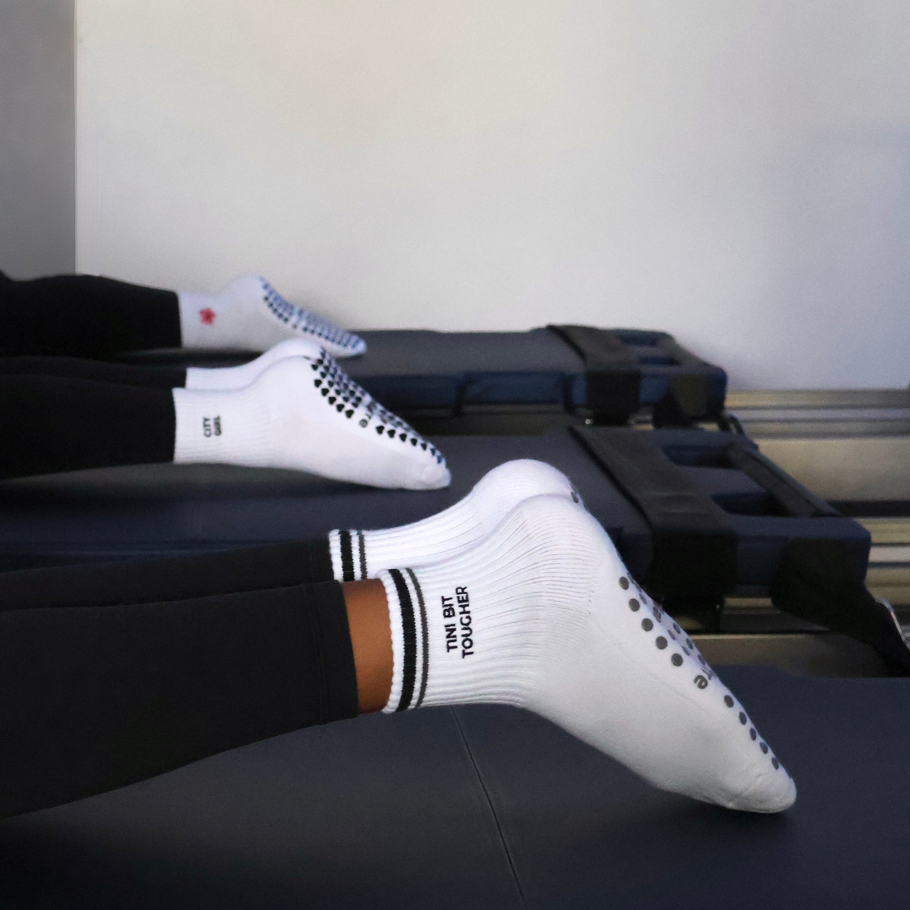 White crew socks with black text and dotted details worn on feet resting on Pilates reformer machines in a studio setting.