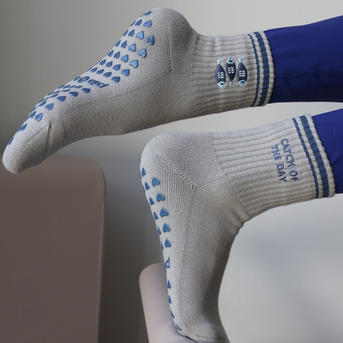 A close-up lifestyle shot of a person's legs resting on a Pilates reformer machine, wearing white grip socks with blue dots and blue leggings.