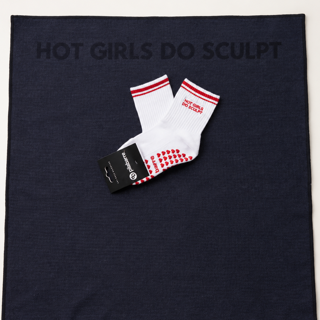 A pair of white crew socks with red stripes and red grip dots, laid flat on a dark navy square cloth that reads 'HOT GIRLS DO SCULPT'. The socks still have a black brand tag attached.