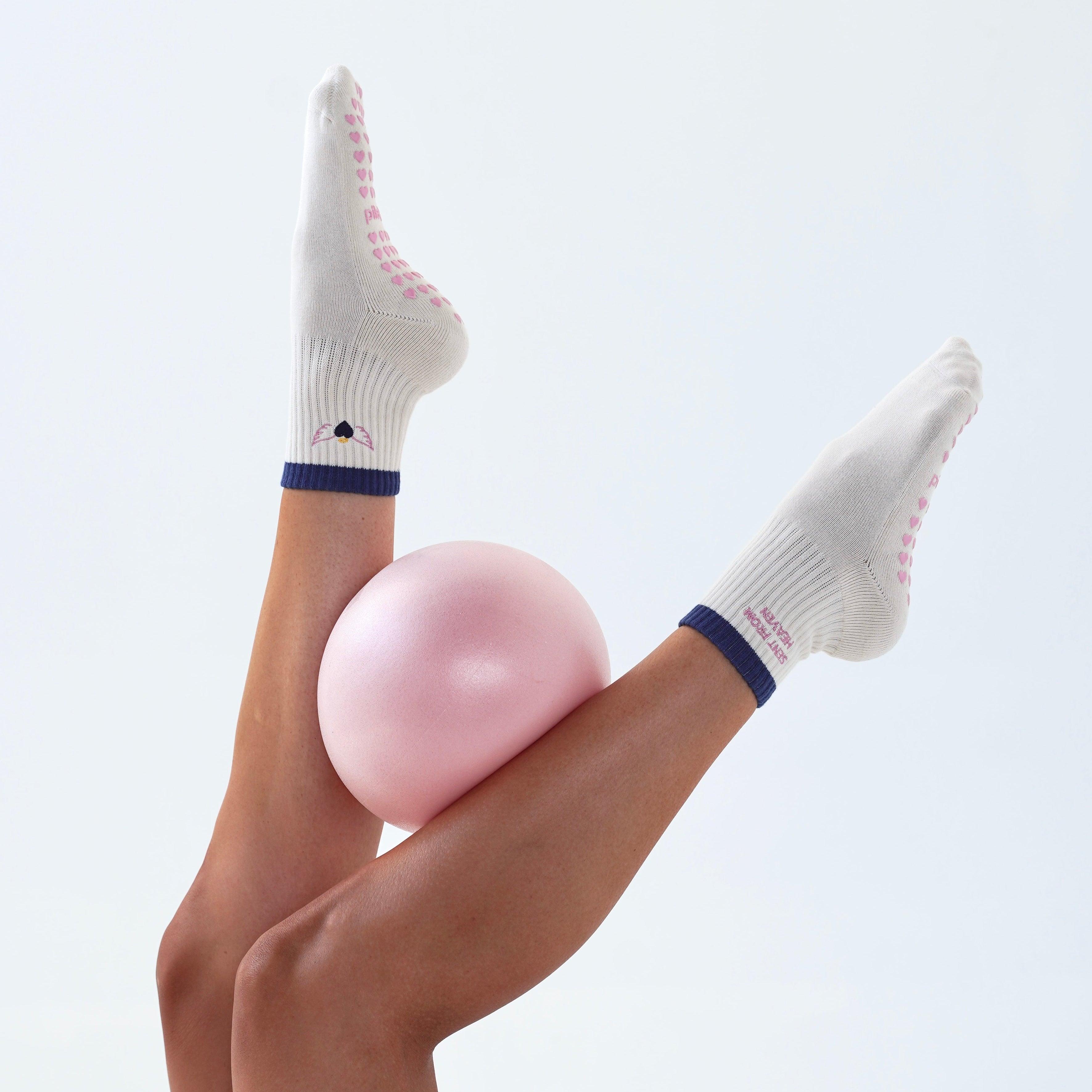 Cream knit grip socks with pink grip dots and navy blue trim, worn on legs extended upward while balancing on a pink exercise ball against a light gray background.