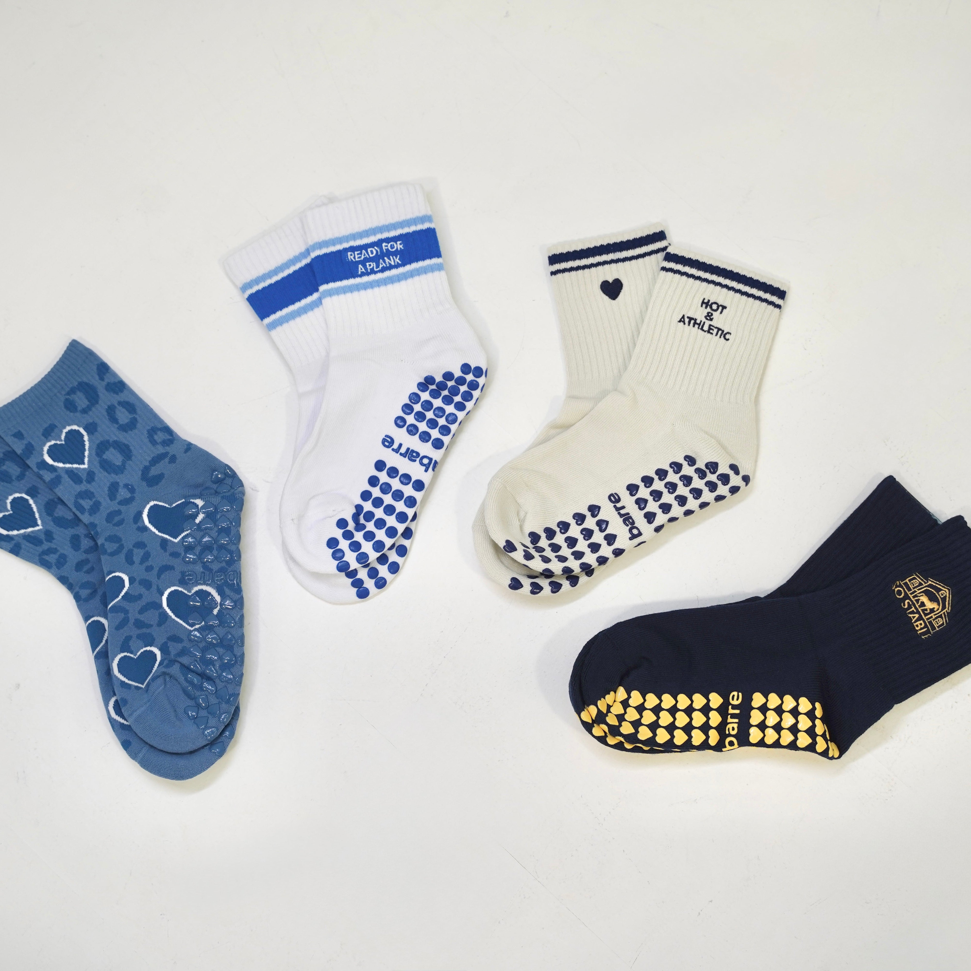Four pairs of grip socks arranged on a plain white background: one blue pair with white heart outlines, one white pair with blue grip dots and text, one cream pair with blue grip dots and a small heart, and one navy pair with gold grip dots and a crest logo. Blue Crush Collection