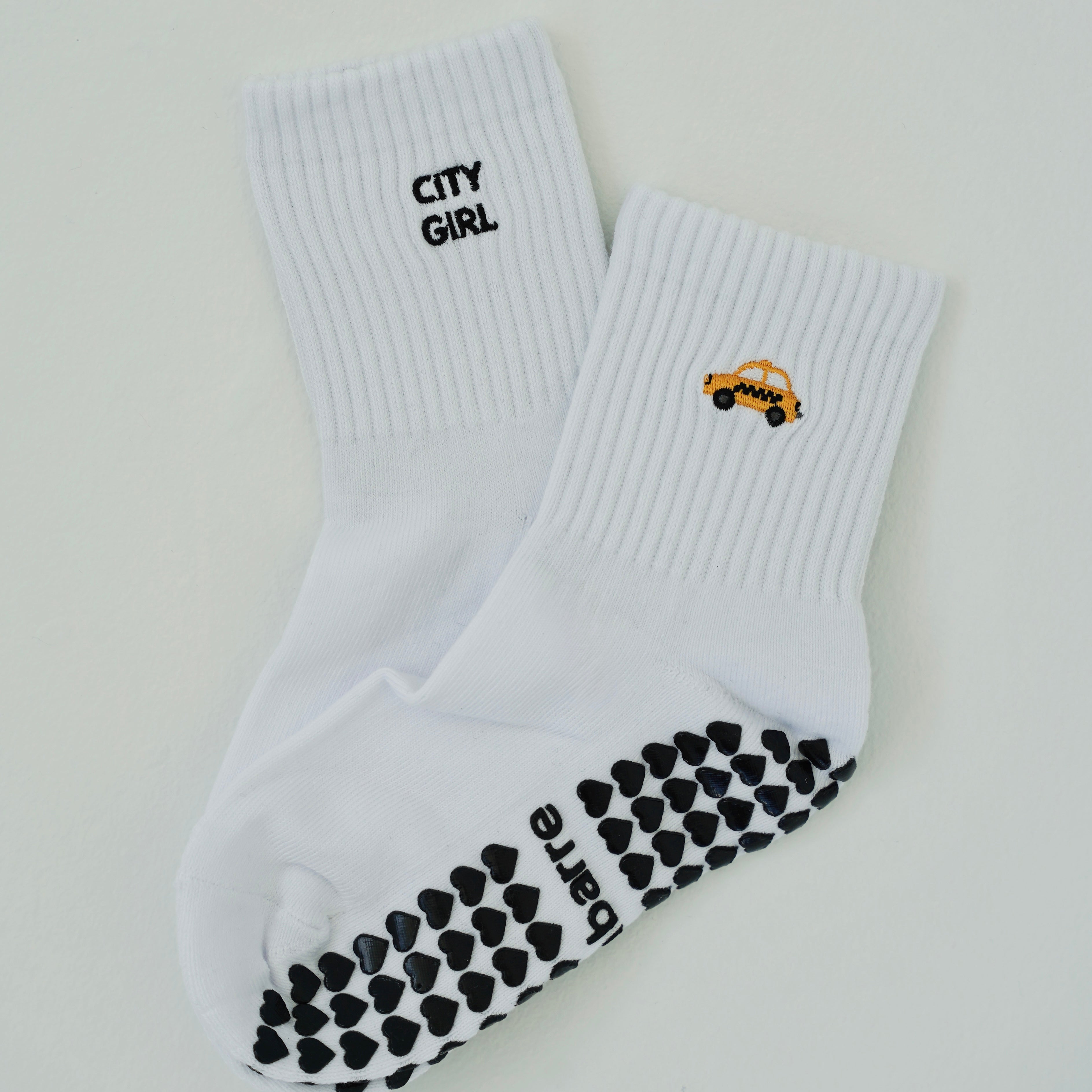 A pair of white crew socks on a light gray background. One sock has 'CITY GIRL' printed in black near the cuff, and the other features a small yellow taxi graphic.