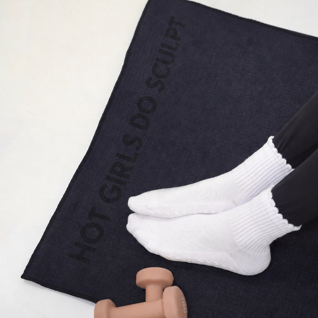 A person wearing white socks and black leggings sits on a black mat towel printed with the text 'HOT GIRLS DO SCULPT'. A small pink dumbbell rests near the bottom of the mat on a light gray floor. HOT GIRLS DO SCULPT BEST EVER PILATES GRIP TOWEL