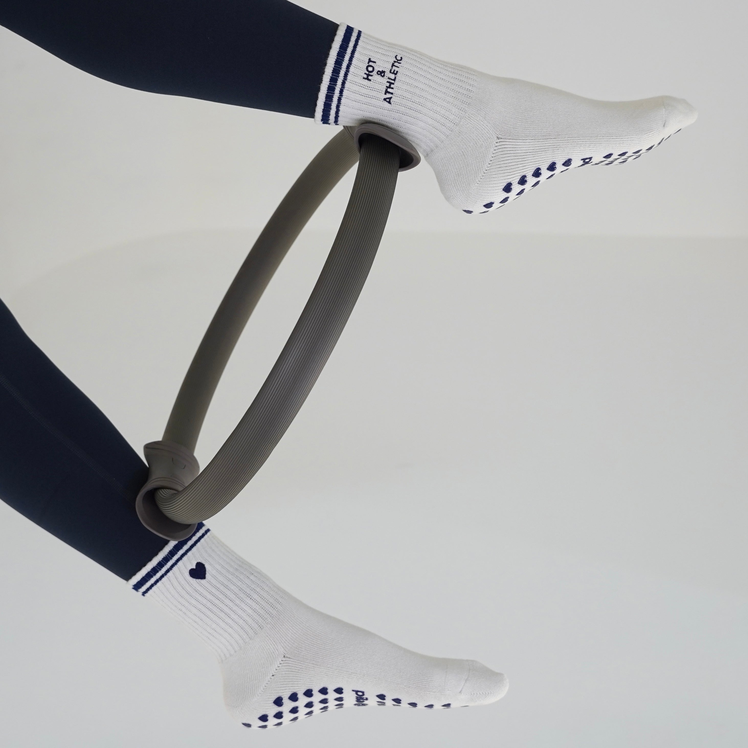 White grip socks with navy blue stripes and grip dots, worn on feet using a grey pilates ring. Hot and athletic grip socks with pilates ring