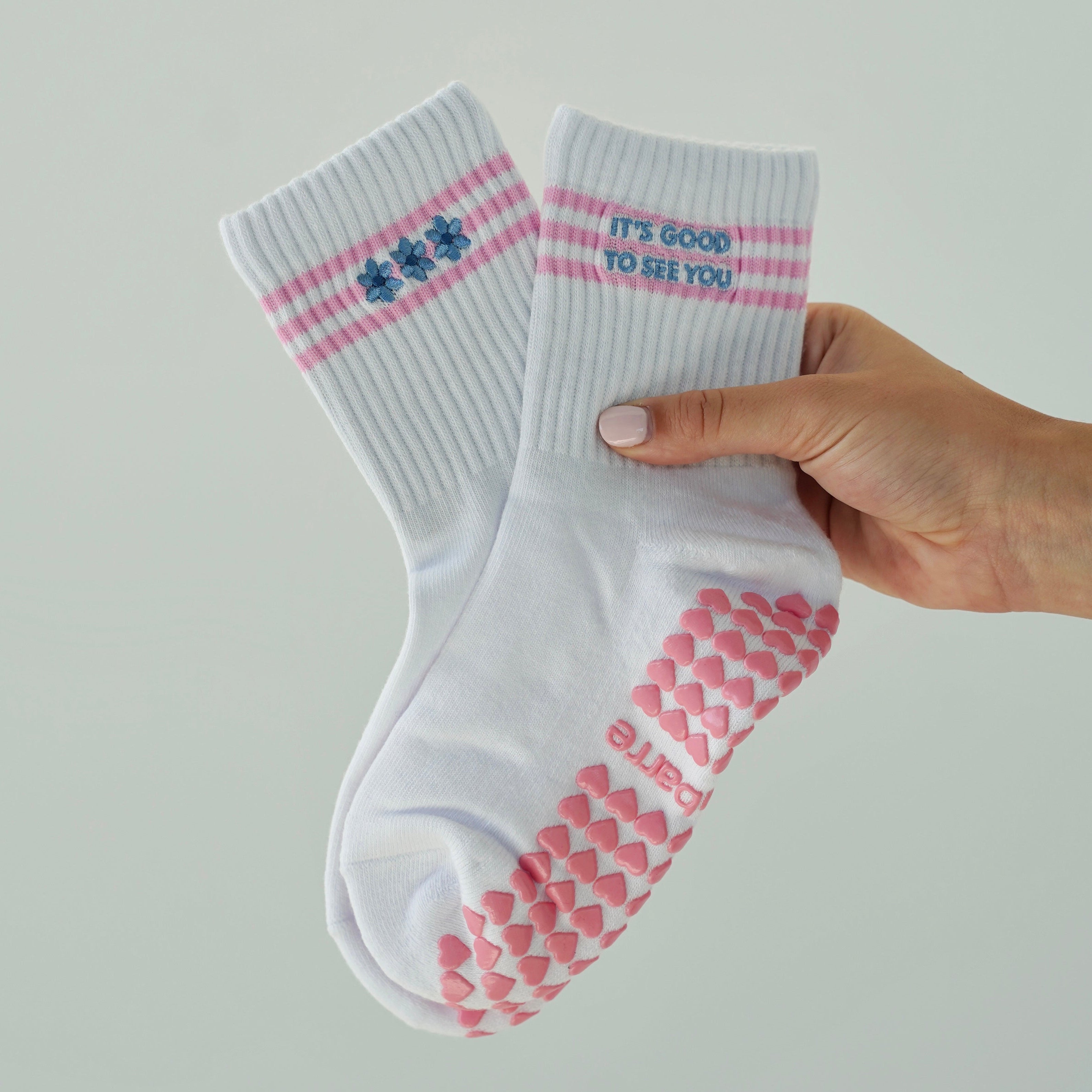 A hand holding a pair of white crew socks with pink grip dots on the soles and pink stripes with blue text on the cuffs, against a light gray background.