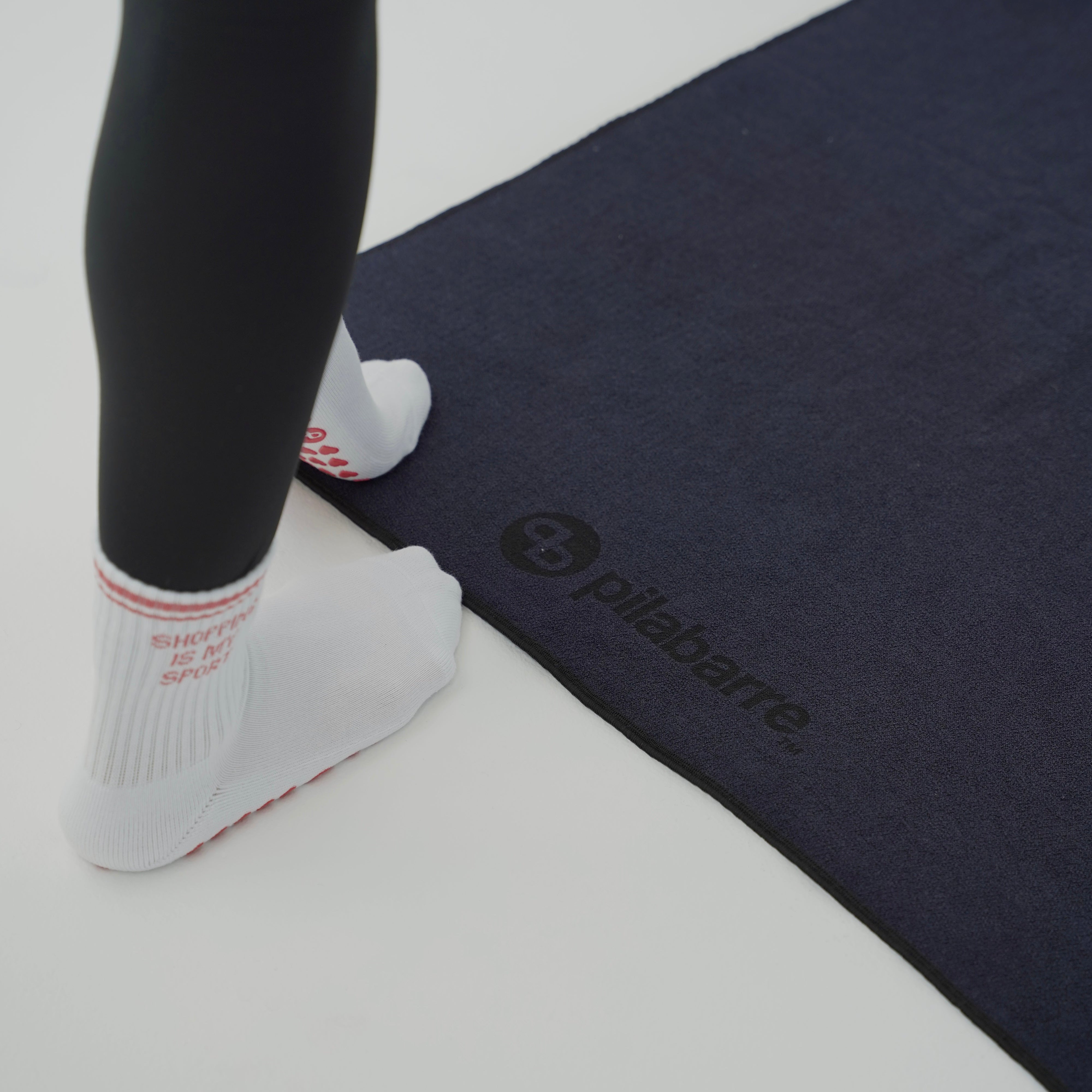A person wearing black leggings and white Pilabarra socks steps onto a dark navy Pilabarra exercise mat on a light gray floor.