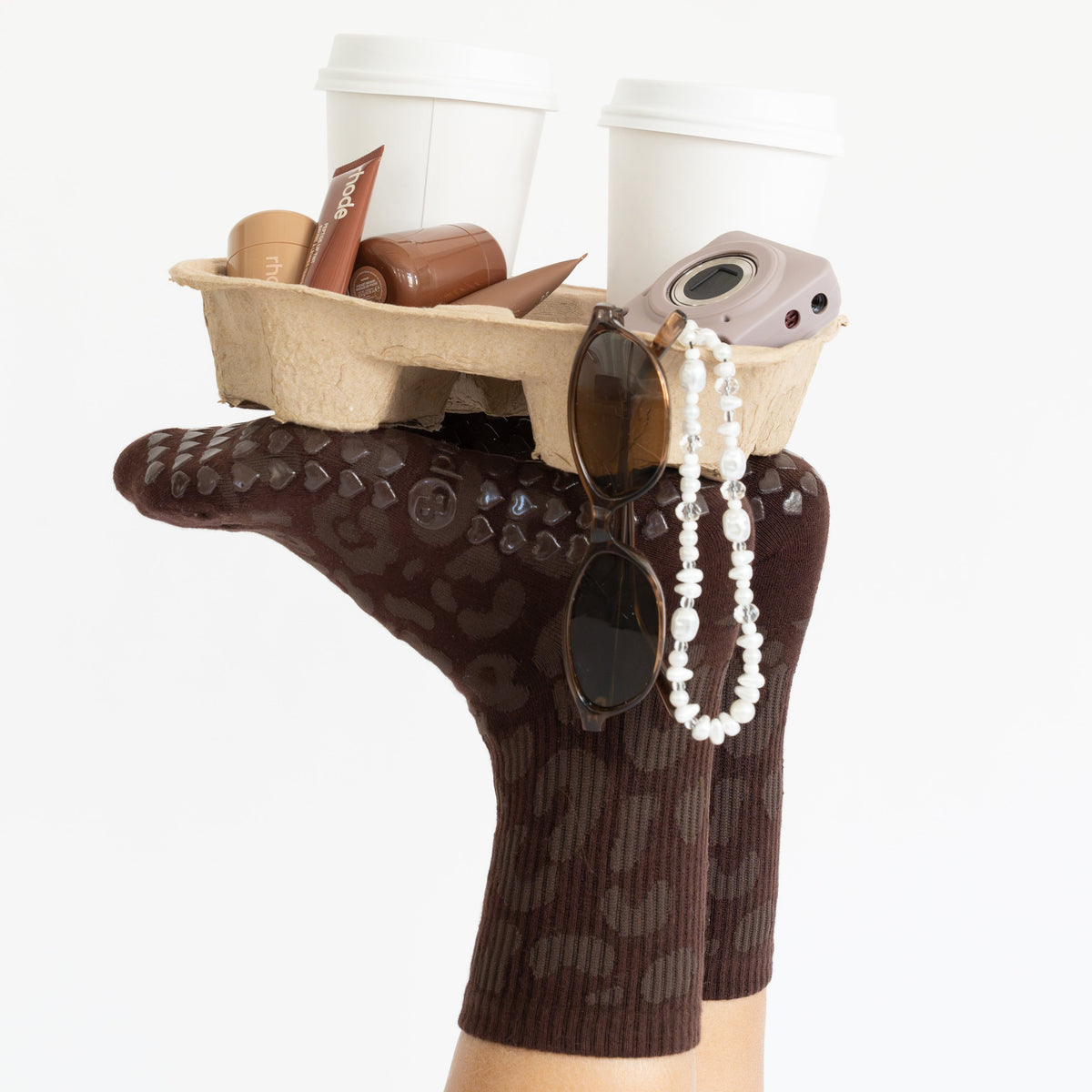 A person's foot wearing a dark brown patterned sock, holding a small cardboard tray with two white takeaway coffee cups, sunglasses, a pearl necklace, and a small camera, against a plain white background.