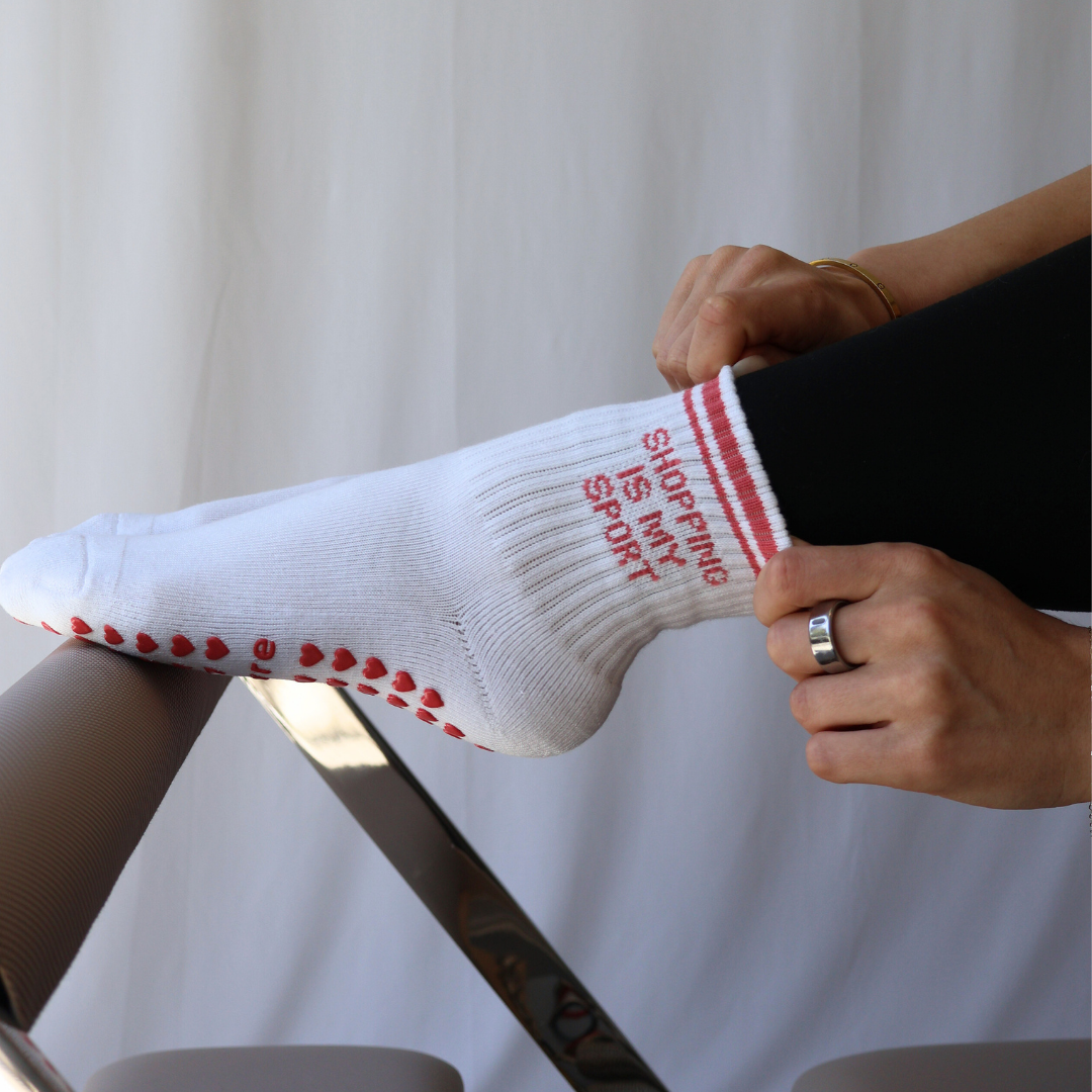 White sock with pink "shopping is my sport" text and heart patterns held by a person against a white curtain background
