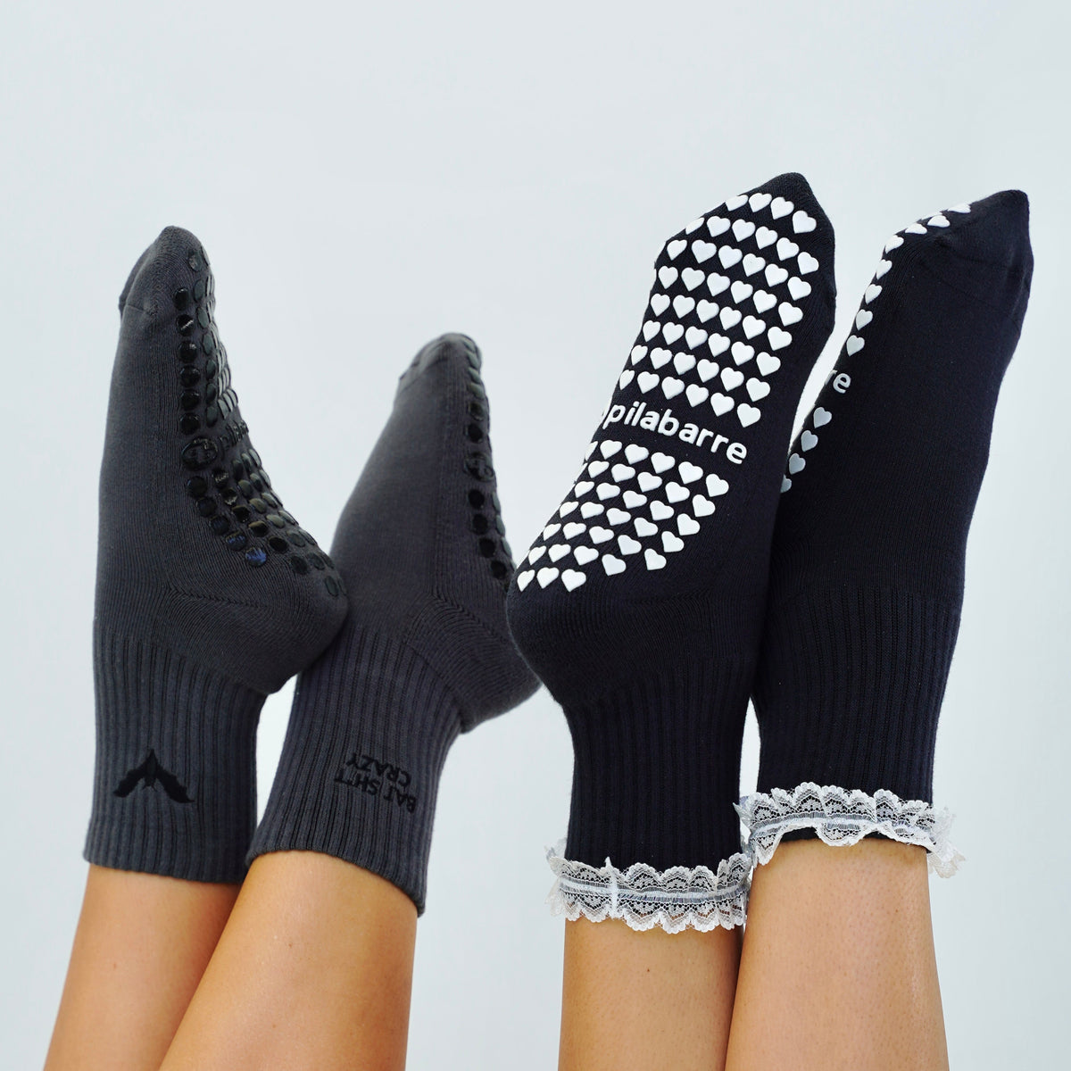 Two pairs of grip socks worn on feet, shown against a plain light background. One pair is dark gray with black grip pads, and the other pair is black with white grip pads and white lace trim.