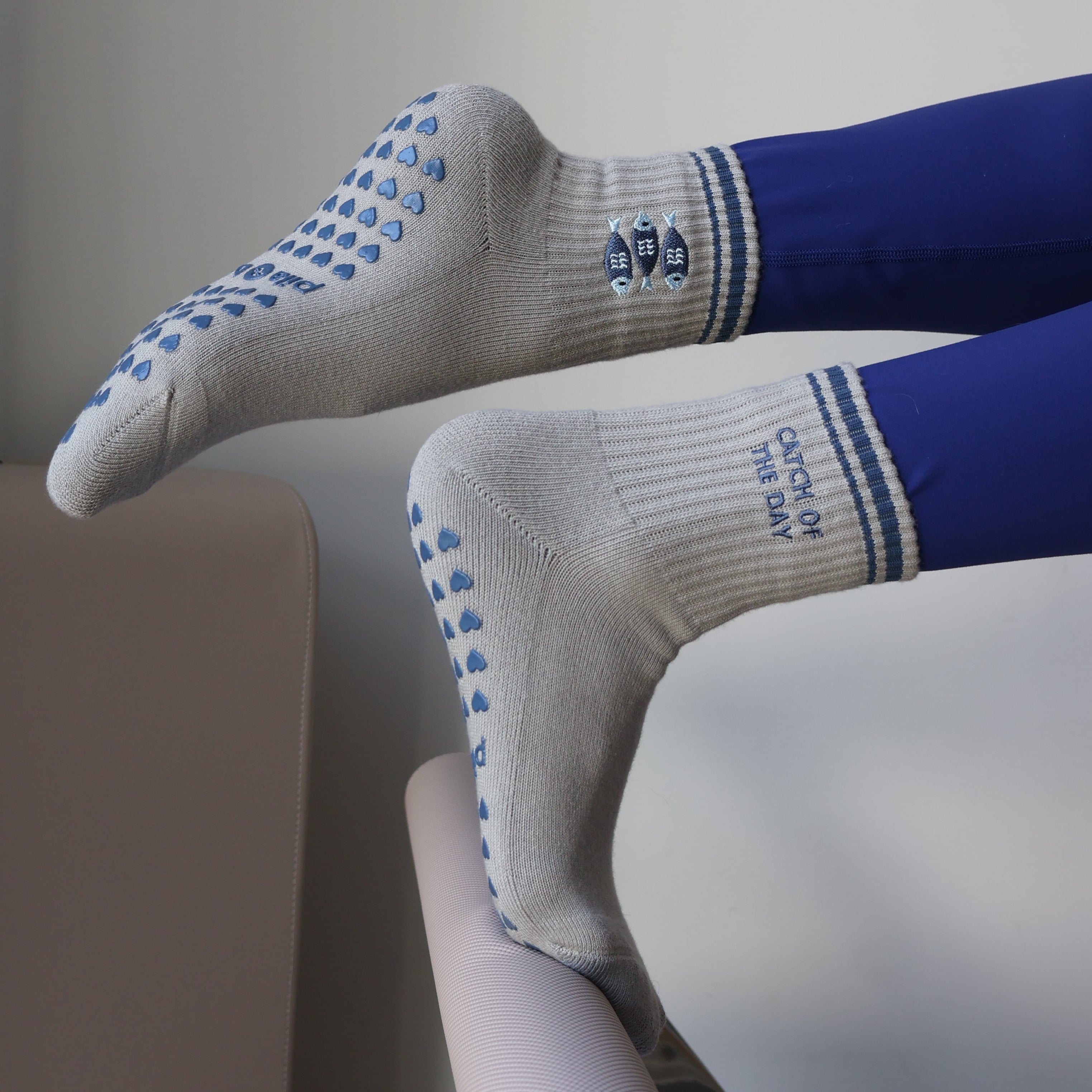 A close-up lifestyle shot of a person's legs resting on a Pilates reformer machine, wearing white grip socks with blue dots and blue leggings.