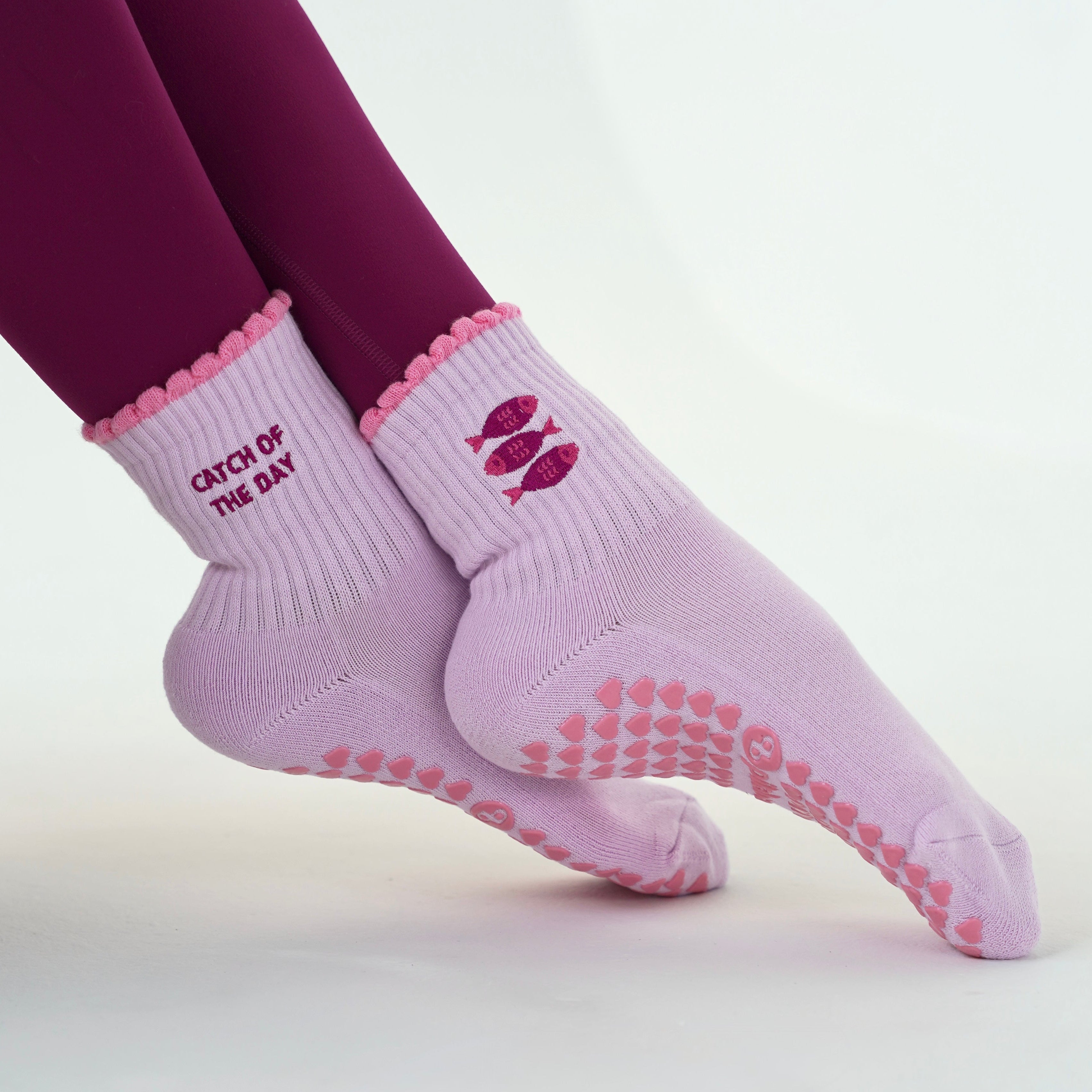 A pair of light pink grip socks with darker pink grip dots on the soles and a lip graphic on the side, worn with dark magenta leggings against a clean white background.