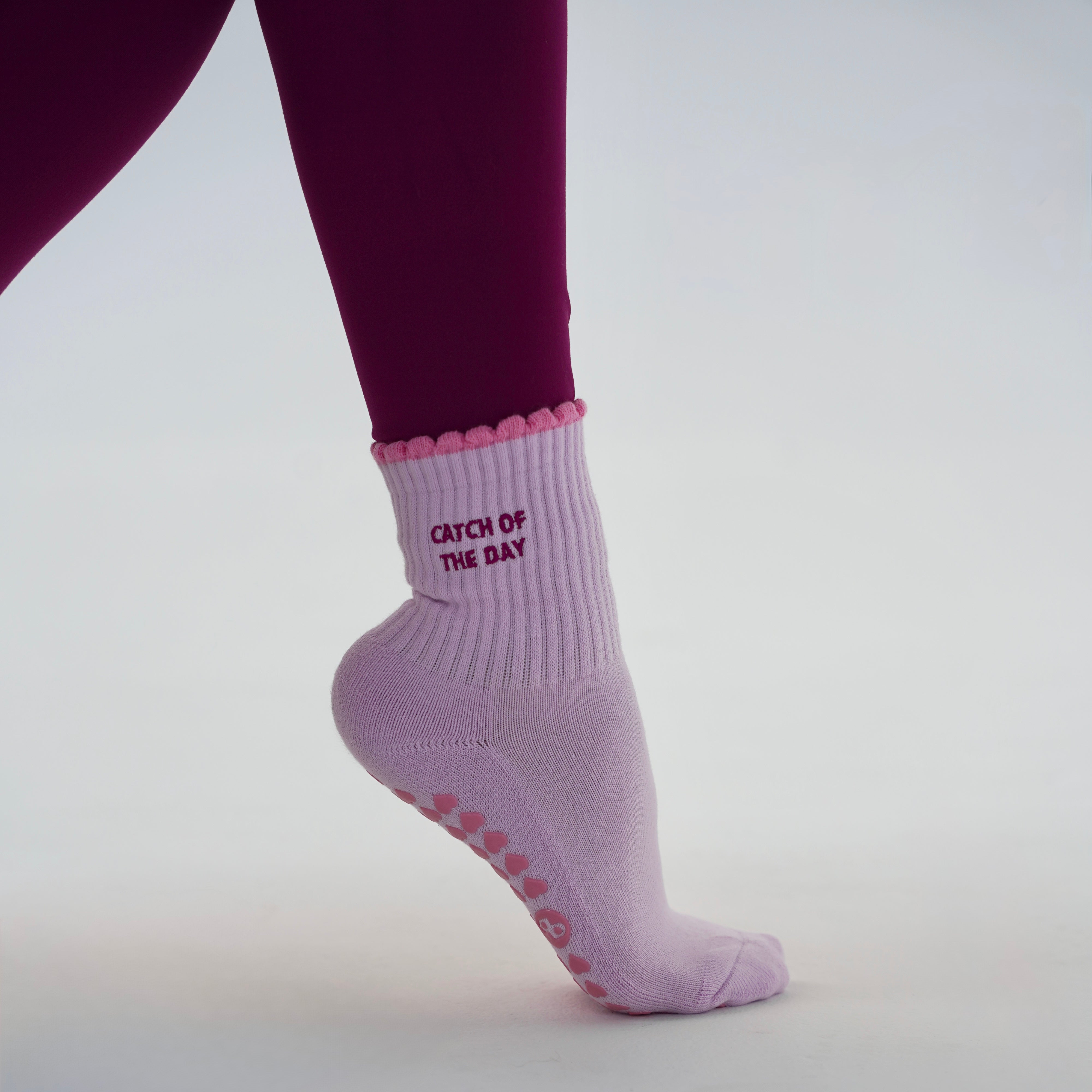 A person wearing a light lavender grip sock with a pink scalloped trim and pink grip dots, standing on a white surface. The sock has 'CATCH OF THE DAY' printed in pink on the side.