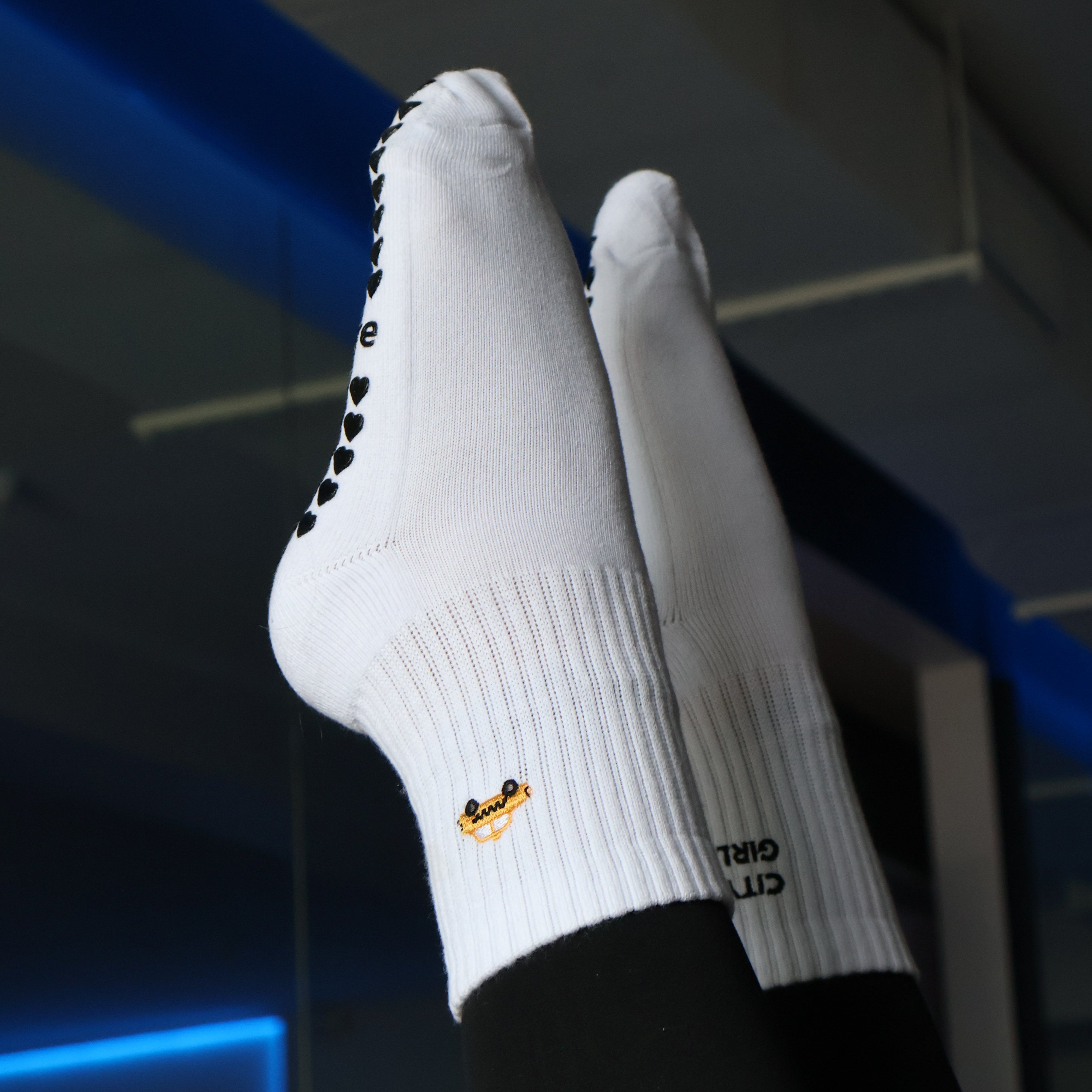 White crew socks with black grip dots on the toes and a small yellow-and-black logo near the cuff, worn on feet raised against an indoor background with blue lighting.
