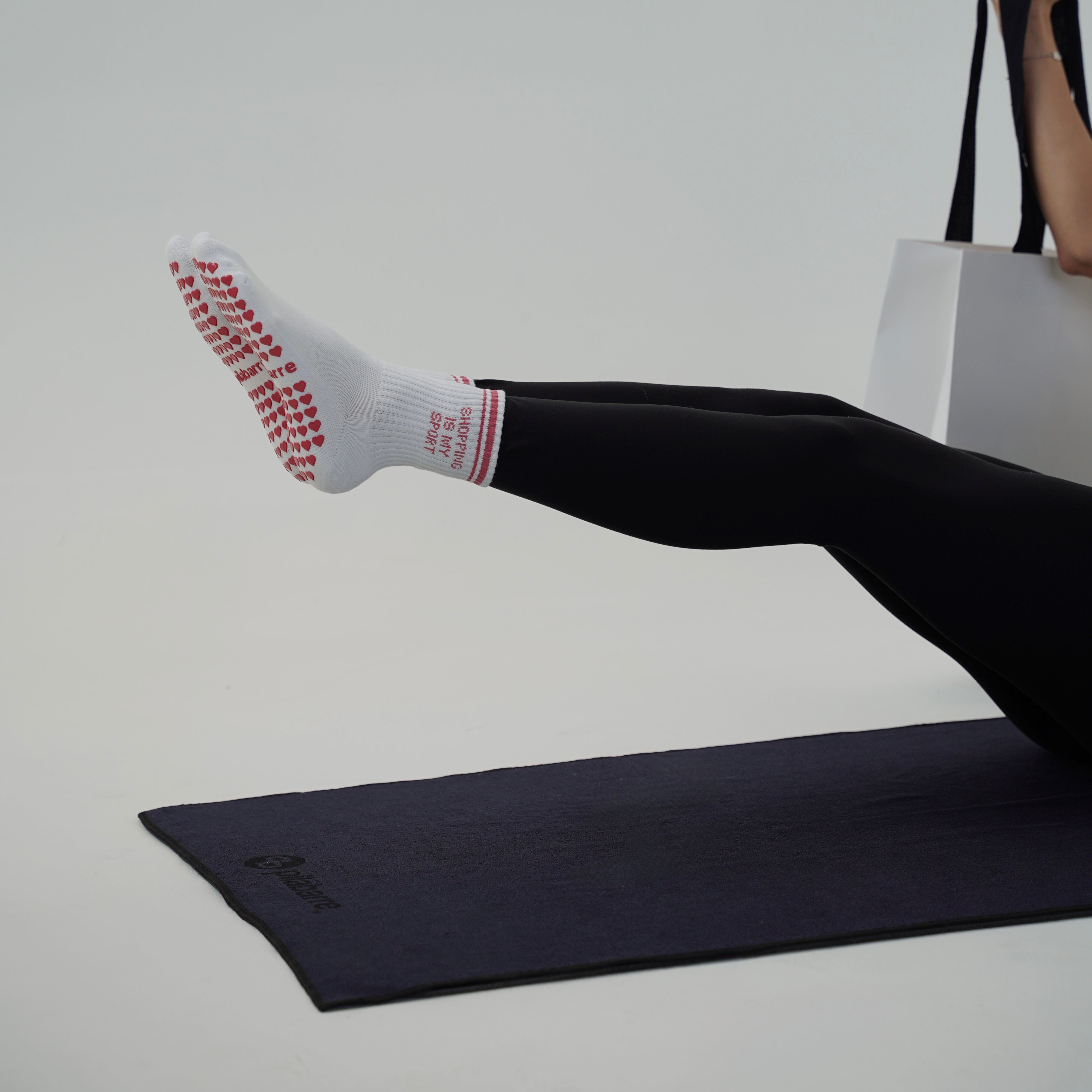 A person in black leggings sits on a dark yoga mat, lifting one leg while wearing a white grip sock with red textured dots and red text. A white tote bag is held in the other hand against a clean light gray studio background.
