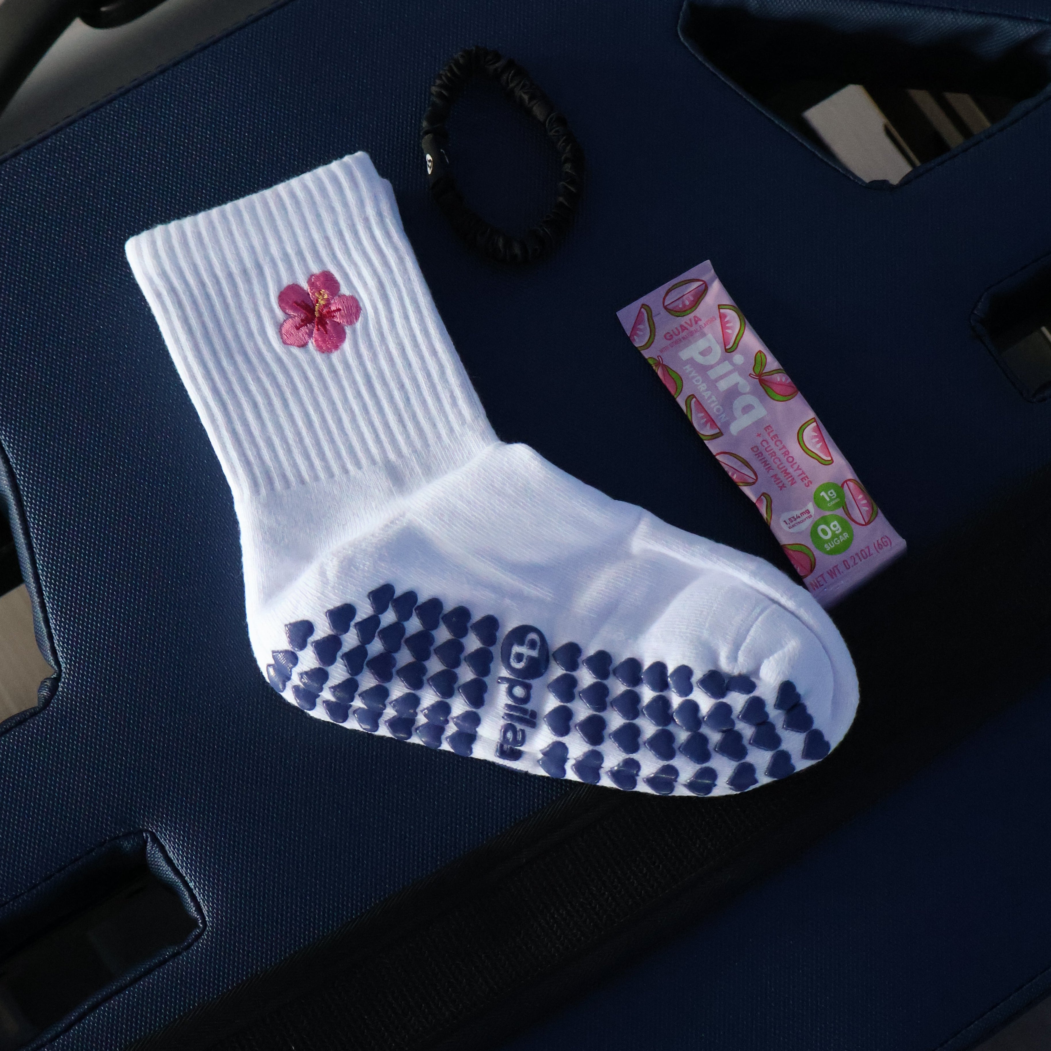 A white ribbed grip sock with a pink hibiscus flower graphic and dark blue heart grips on the sole, displayed on a dark blue reformer alongside a black hair tie