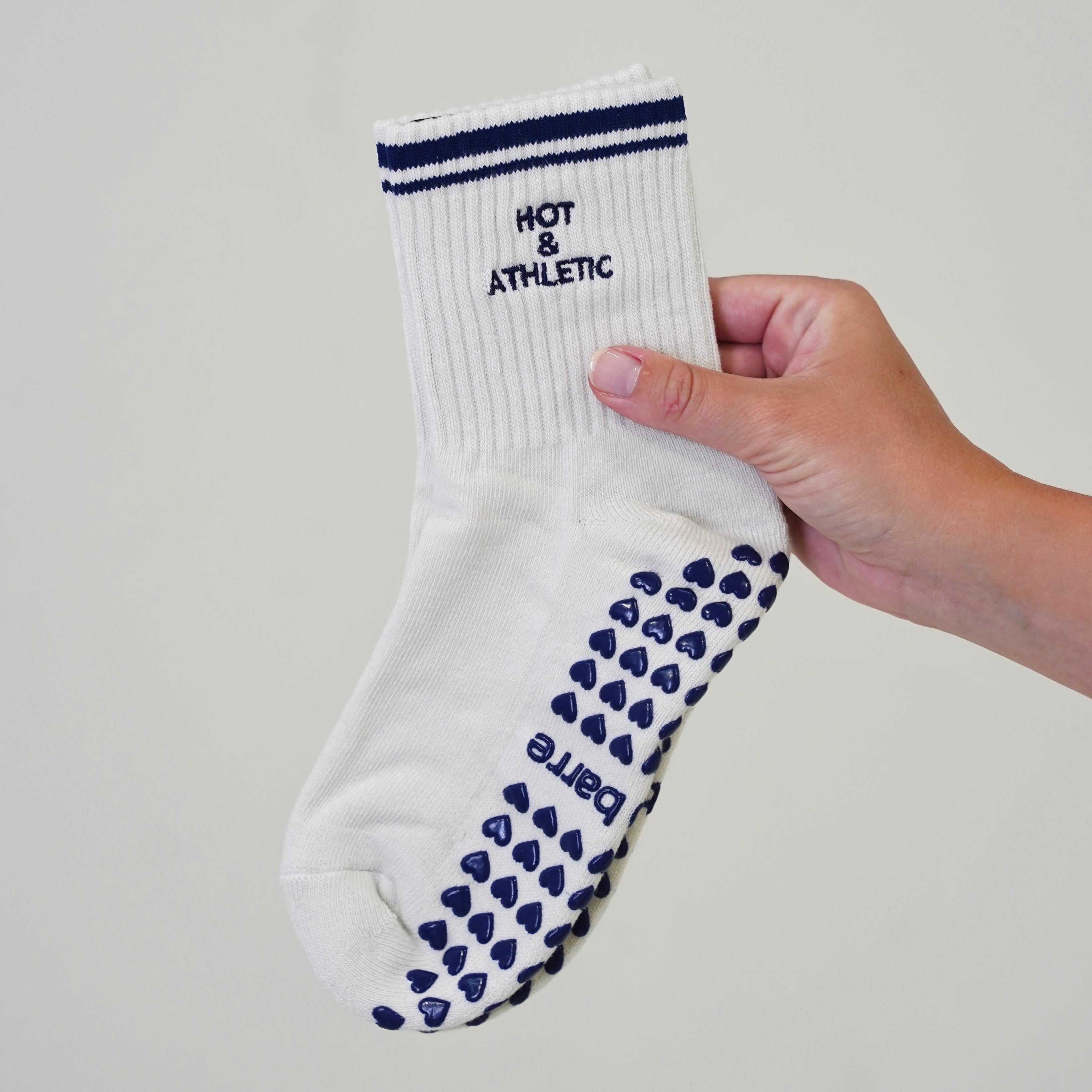 A hand holding a white ribbed sock with navy blue stripes at the cuff and navy blue grip dots on the sole, featuring the text 'HOT & ATHLETIC'. hot and athletic barre socks