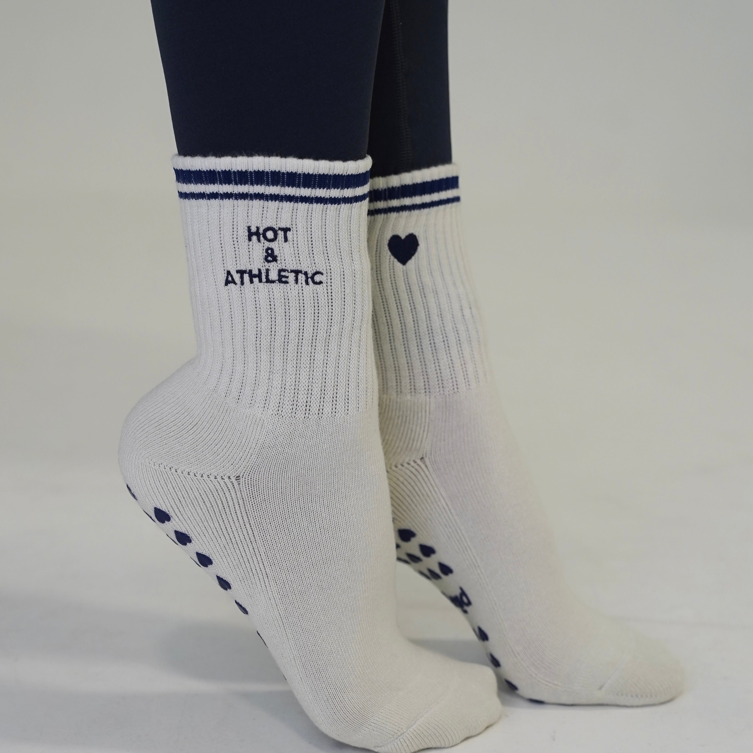 Off white athletic crew socks with navy blue stripes, the words 'HOT & ATHLETIC' on one sock, a small heart on the other, and navy grip dots on the soles, shown on a person's feet against a light gray background. hot and athletic grip socks - cute pilates socks