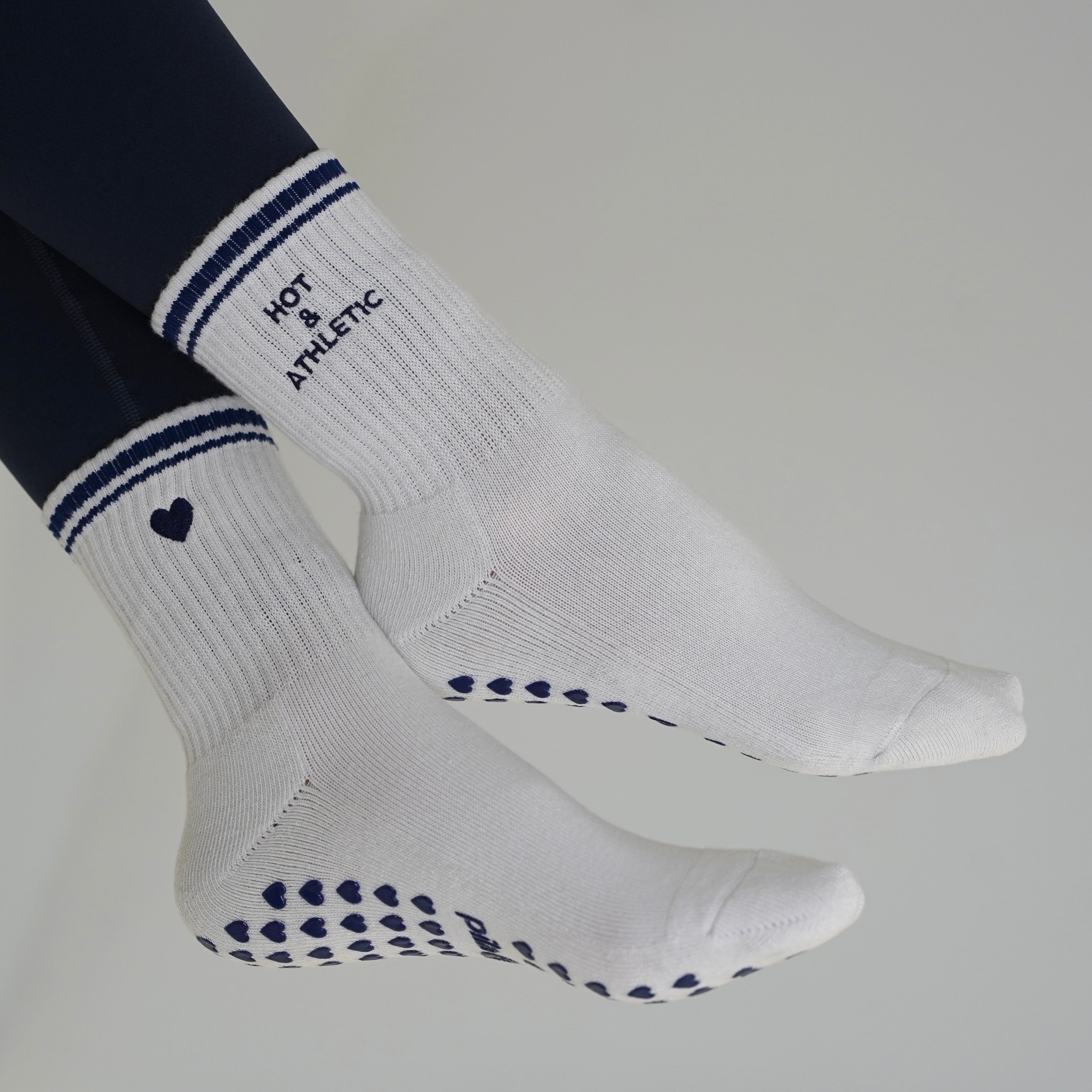 off white athletic crew socks with navy blue stripes, a small heart logo, and grip dots on the sole, worn with navy leggings against a light gray background. hot and athletic grip socks from pilabarre