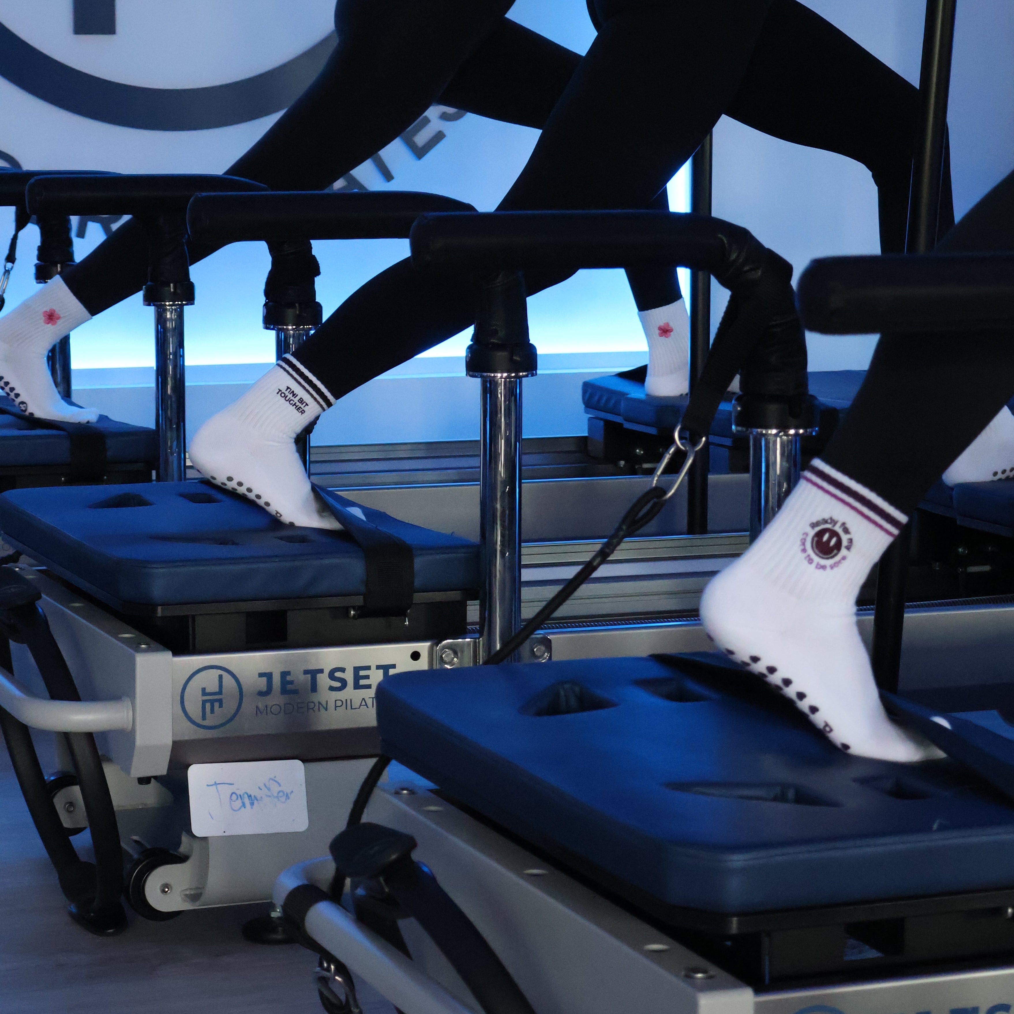 Two women in black activewear and white socks perform Pilates exercises on Jetset Modern Pilates reformer machines in a gym setting.