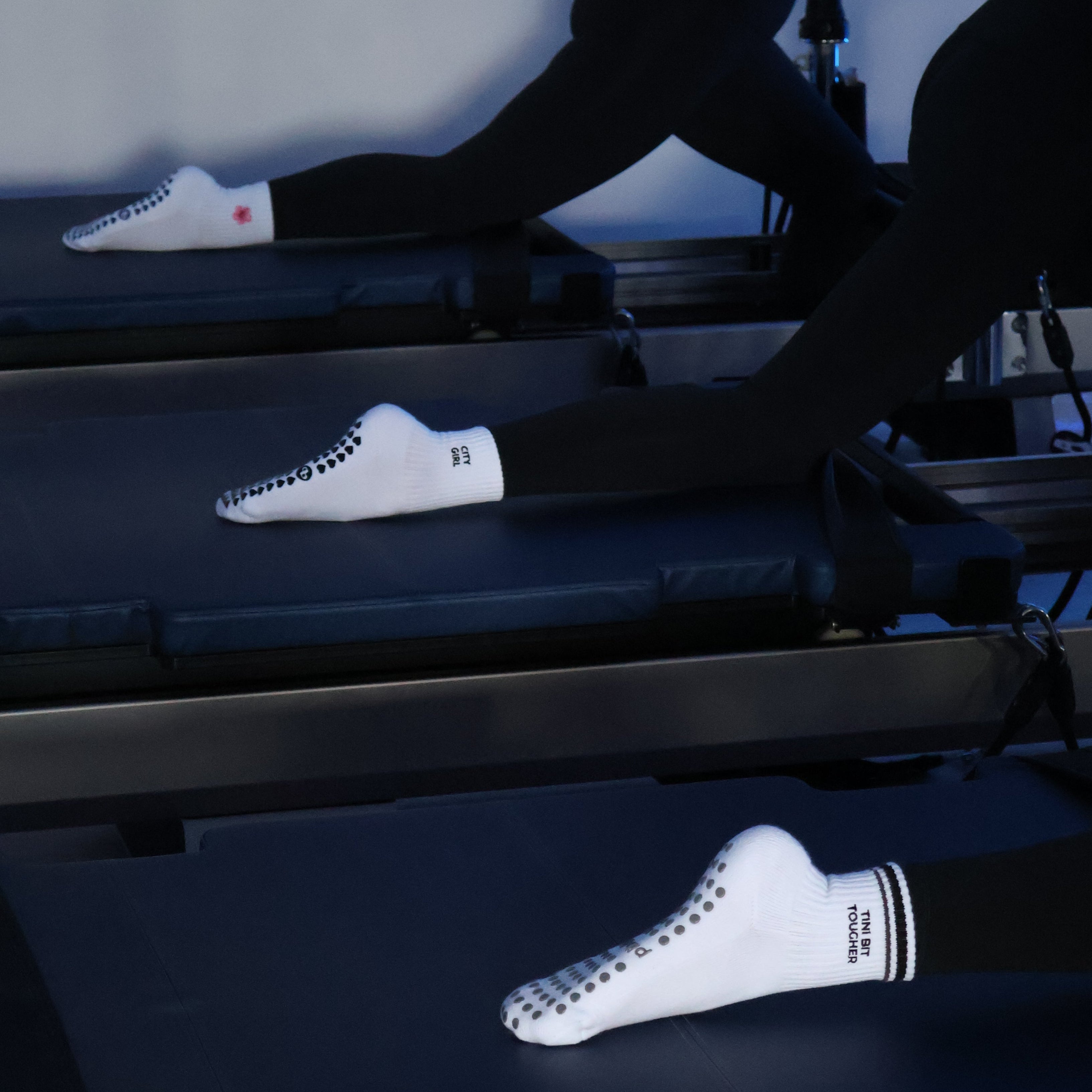Two women in black activewear and white socks perform a pilates exercise on reformer machines in a studio setting.