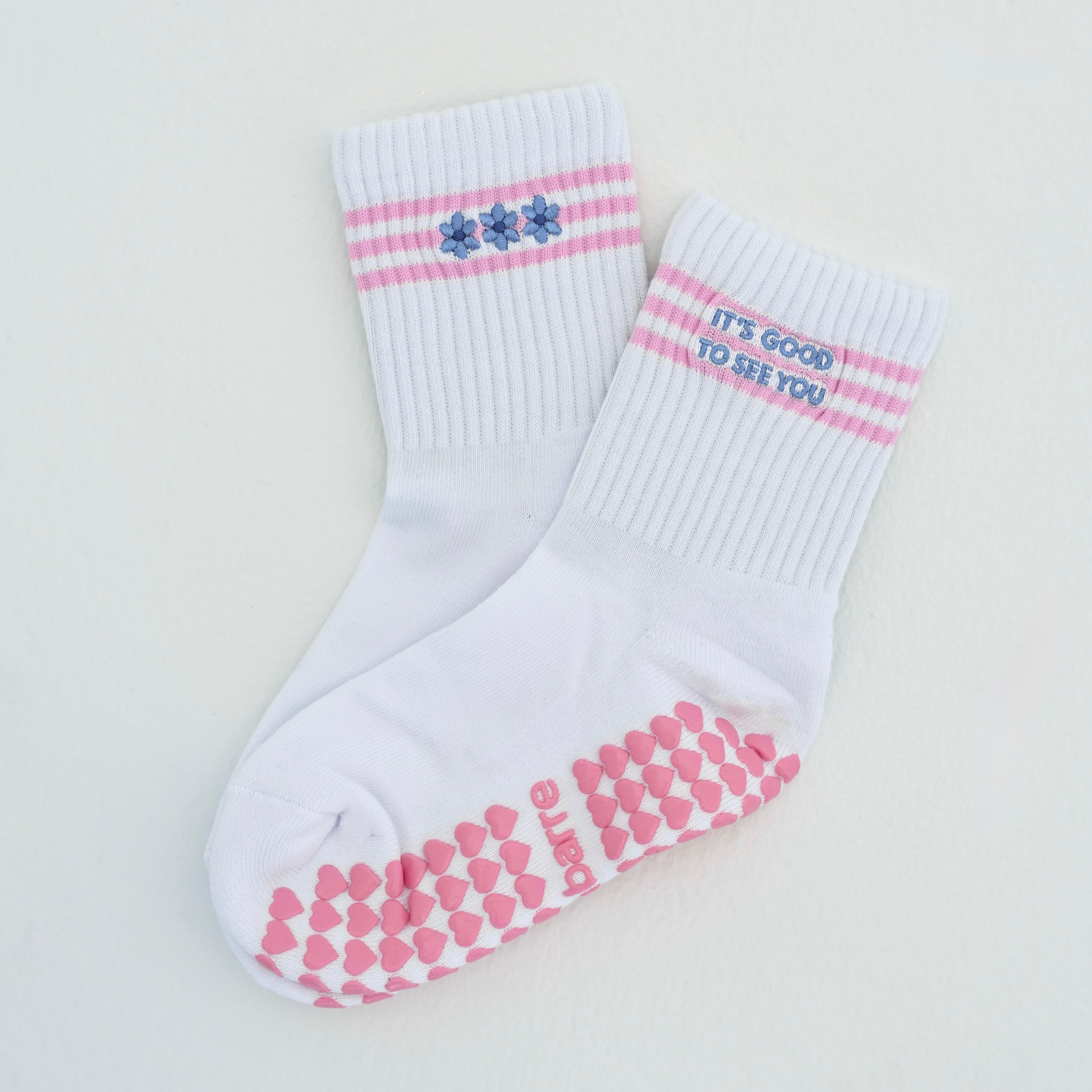 A pair of white crew socks with pink stripes, blue flowers on one cuff, and 'IT'S GOOD TO SEE YOU' text on the other cuff, plus pink grips on the soles.