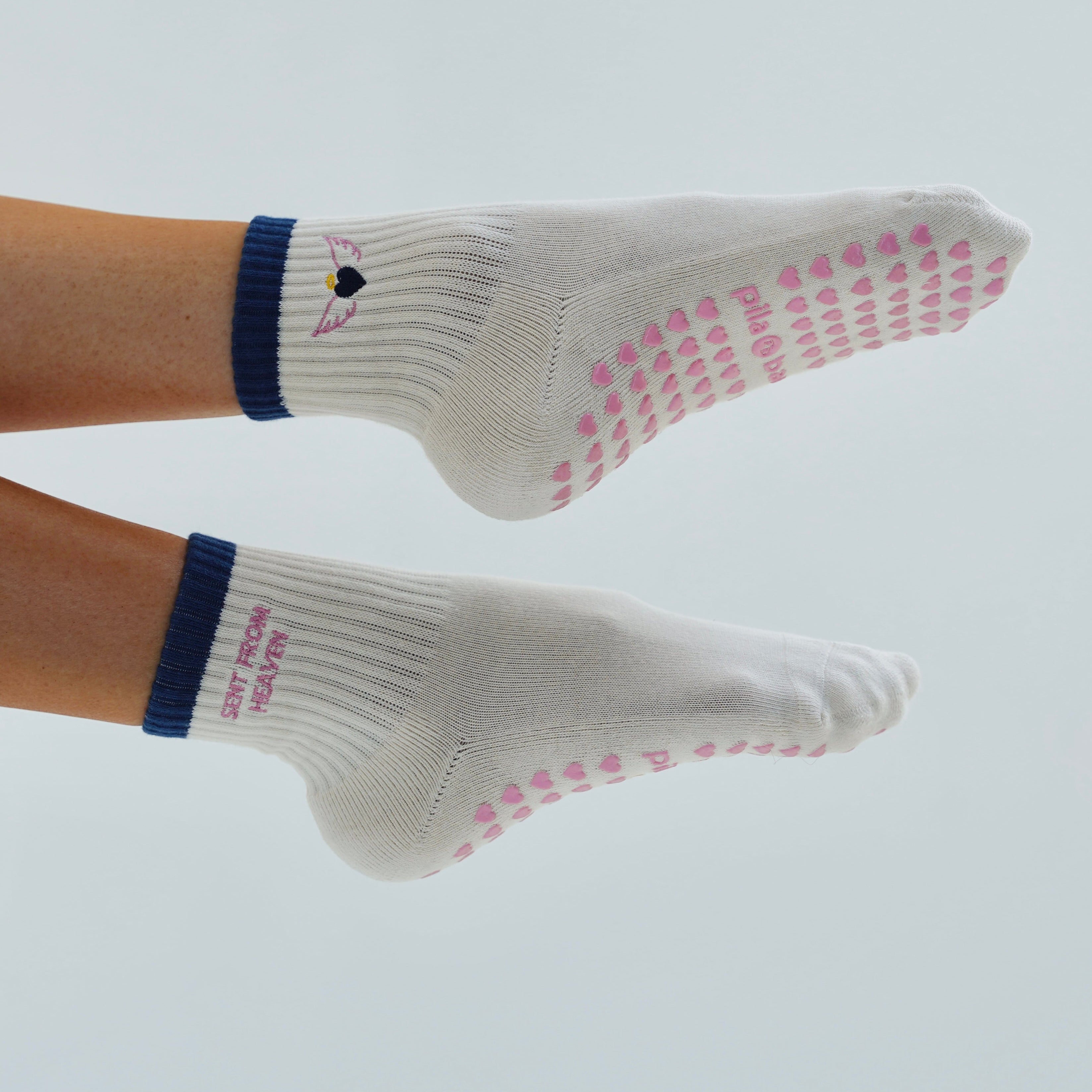 Cream grip socks with pink heart-patterned non-slip soles and navy blue ribbed cuffs, shown on a person's feet against a light gray background.