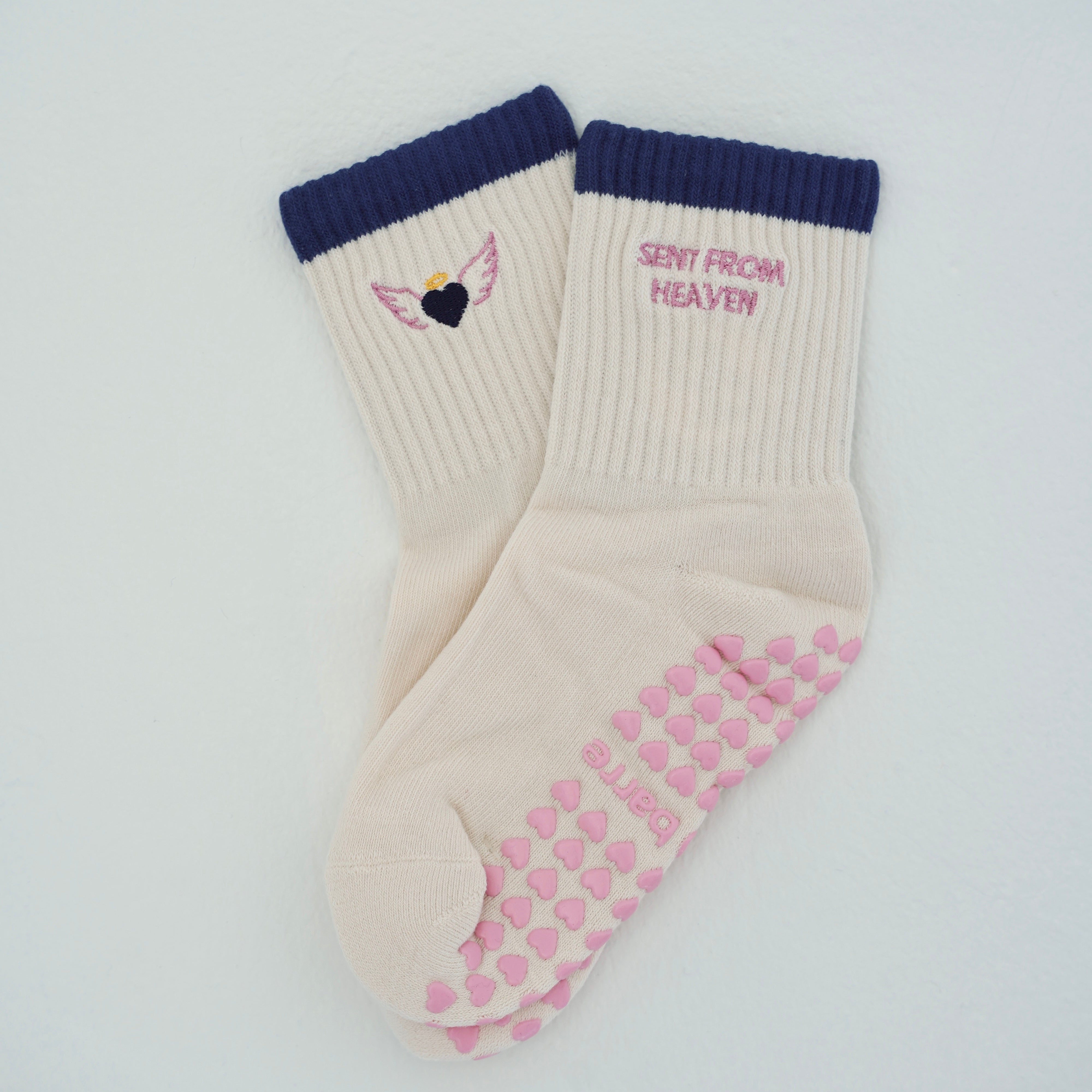 A pair of cream-colored ribbed crew socks with navy blue cuffs. One sock features a small pink and yellow angel wing graphic with a navy heart, and the other features pink text reading 'SENT FROM HEAVEN' above a pink grip pattern on the sole.