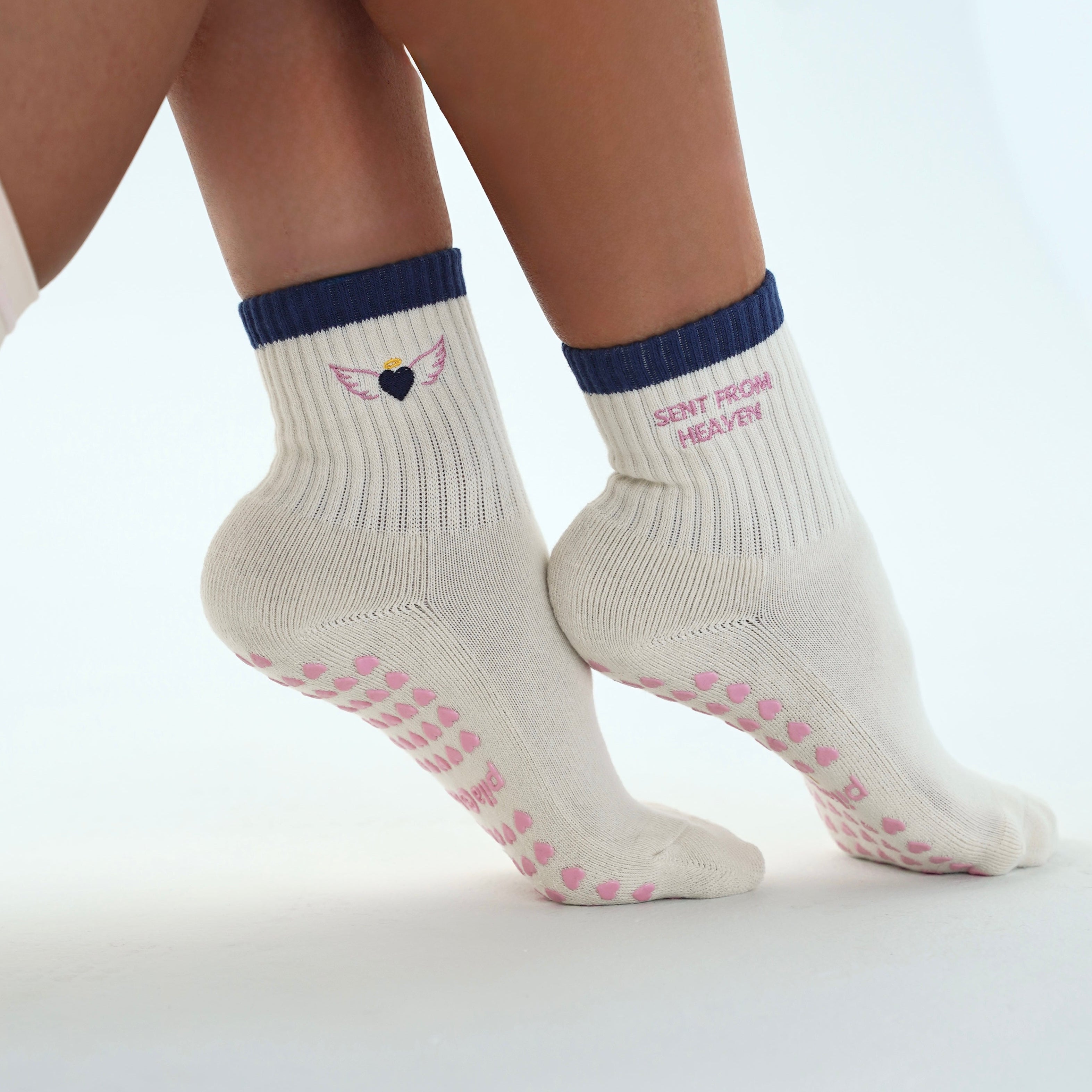 A close-up of a person's feet wearing off-white grip socks with navy blue cuffs, pink grip dots, and a small winged heart logo on the side. Sent from heaven is embroidered on the grip socks