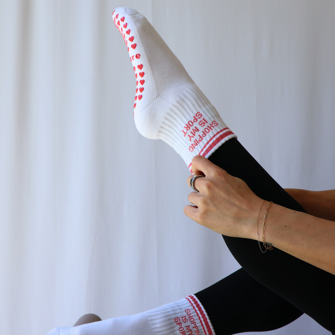 White socks with pink "shopping is my sport" text and heart patterns worn by a person against a white background